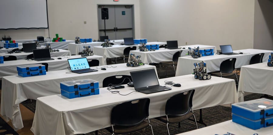 Empty workshop room with VEX IQ kits, robots, and laptop on each table.