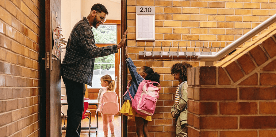 A male teacher stands at a classroom door high-fiving young students as they enter.