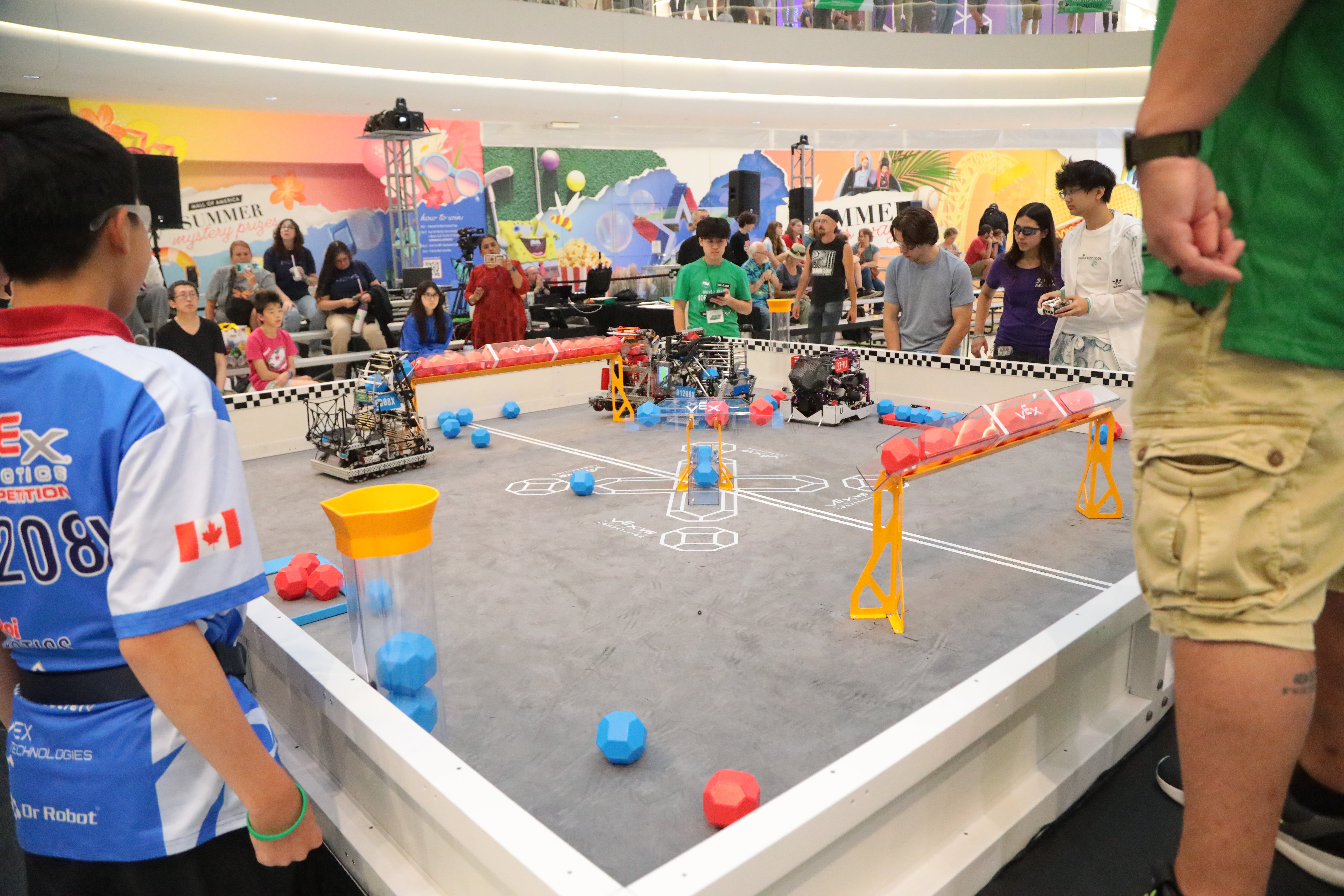 Students in safety glasses line two sides of a v5 field during a match. Four robots can be seen on the field. A scorekeeper stands on the far side with a tablet.