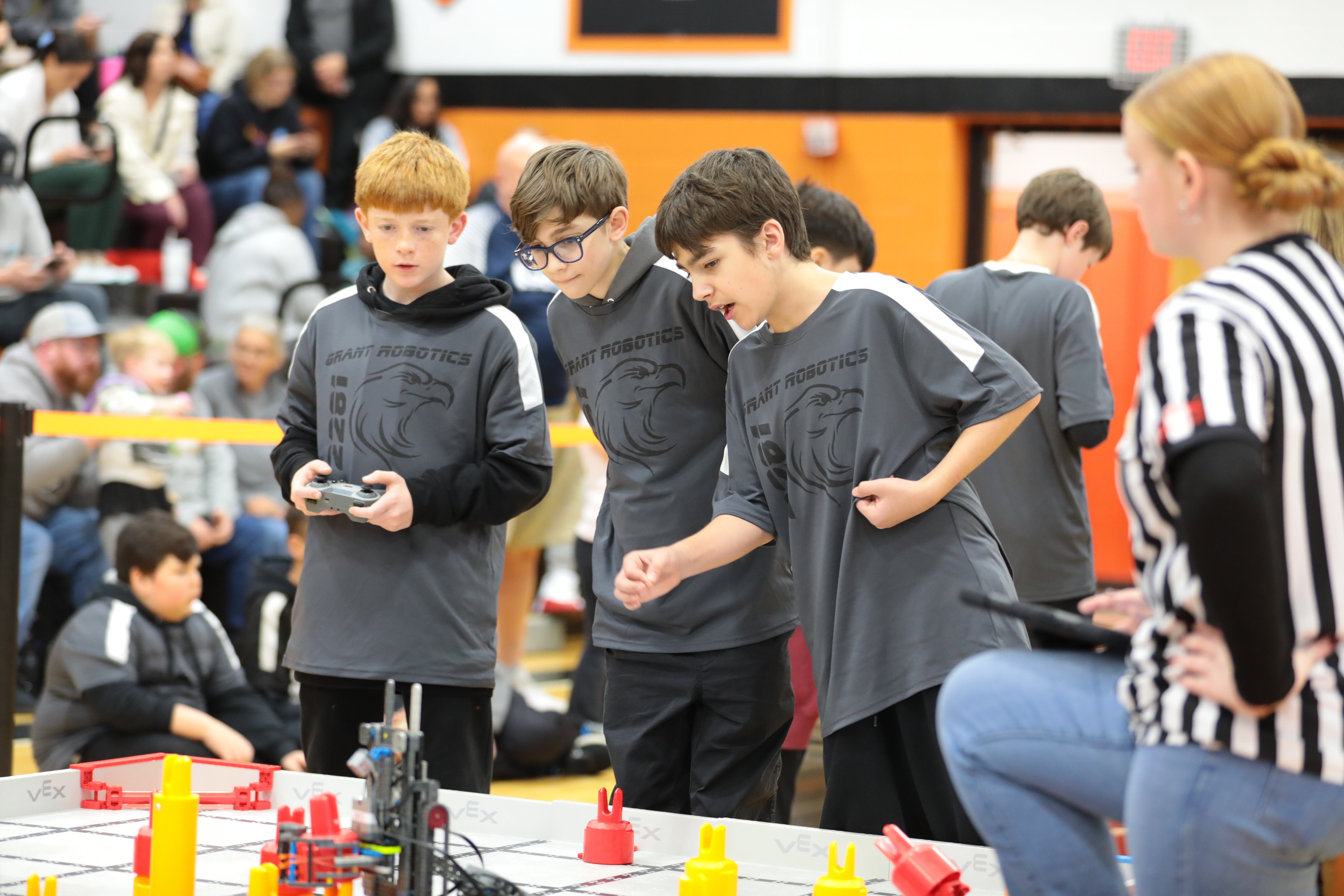 Three students in matching gray team shirts stand at the edge of a VEX IQ field during a match. One holds the controller while another leans forward to watch the robot. A referee in a black-and-white striped shirt watches from the side of the field, and spectators fill the bleachers behind them.