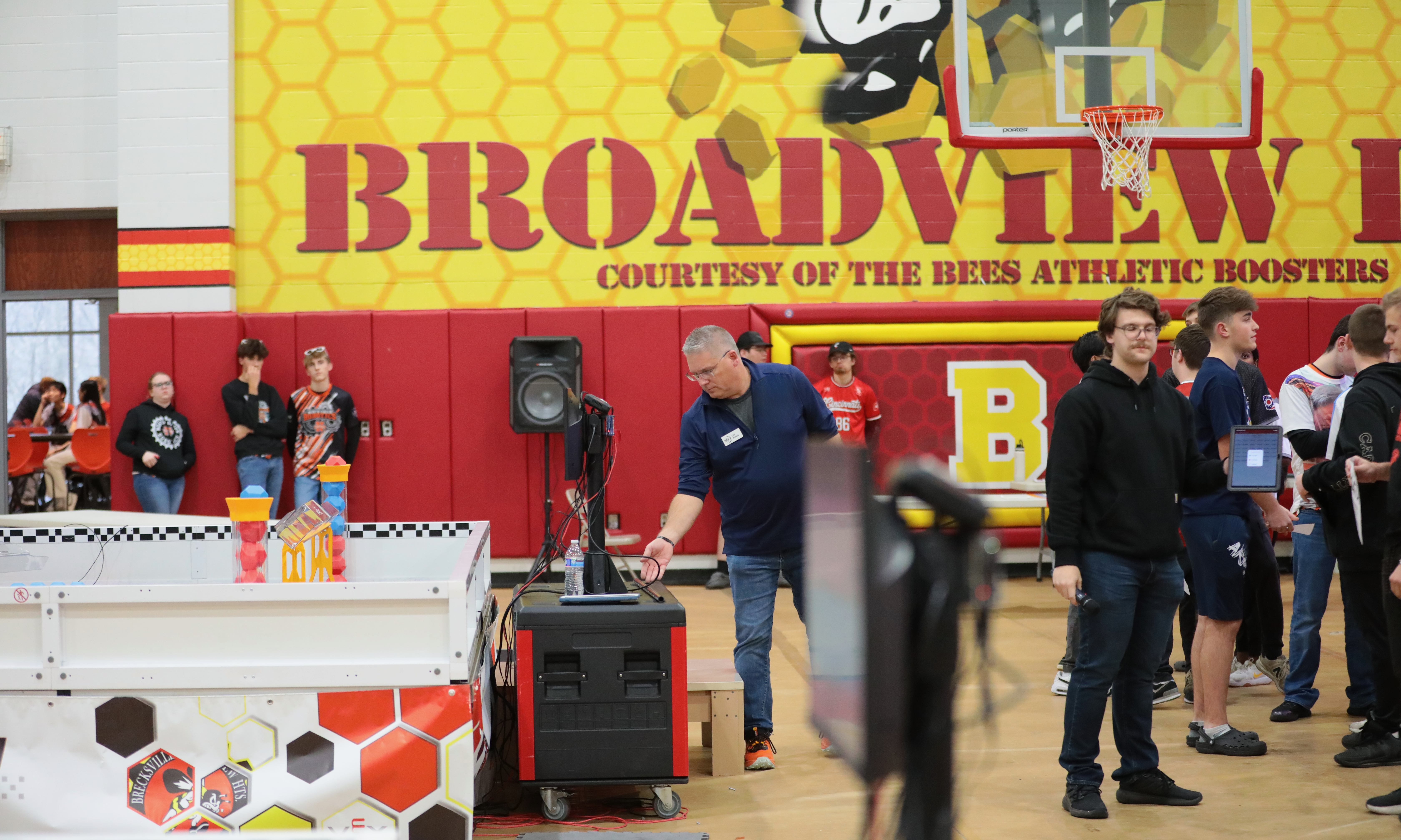 Volunteers in various roles inside a gym at a competition. One is checking technology beside a field, another is queueing, and others are around the outskirts working with teams.