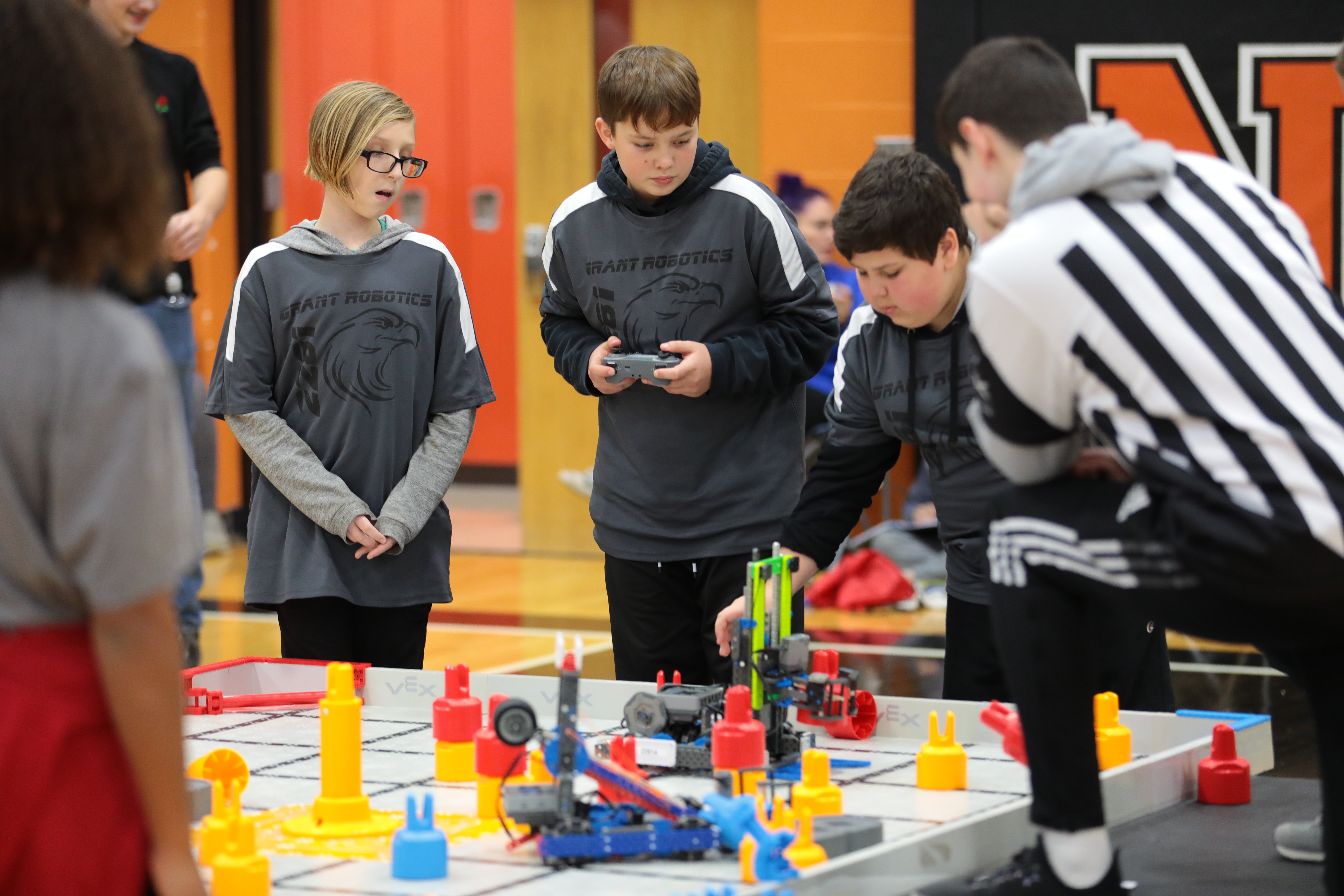 A referee in a striped shirt bends over a VEX IQ field to look closely at the robots. Three students in gray team shirts stand on the other side of the field holding their controllers.