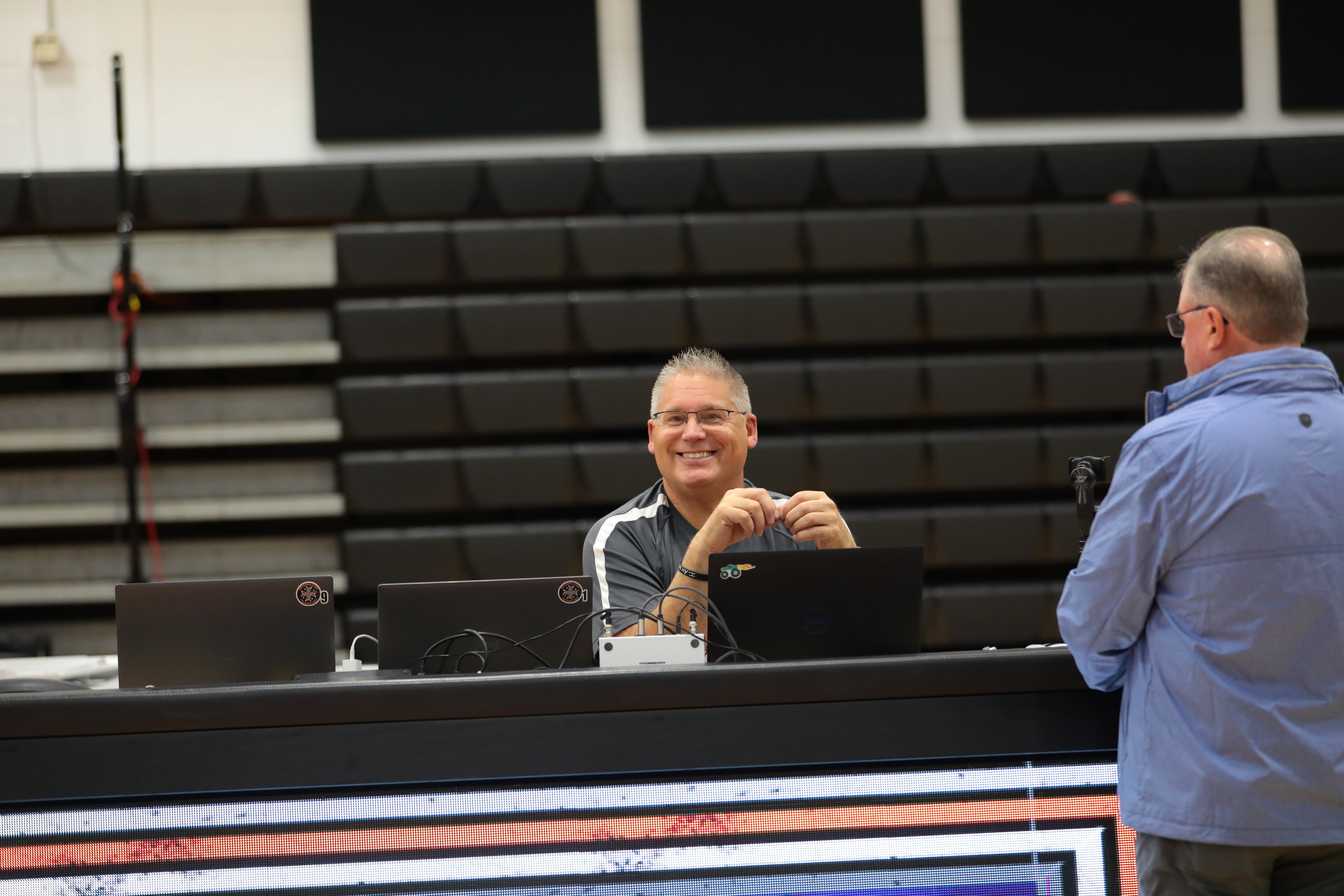 A volunteer sits at a table behind a series of laptops smiling at the camera as he prepares for an event.