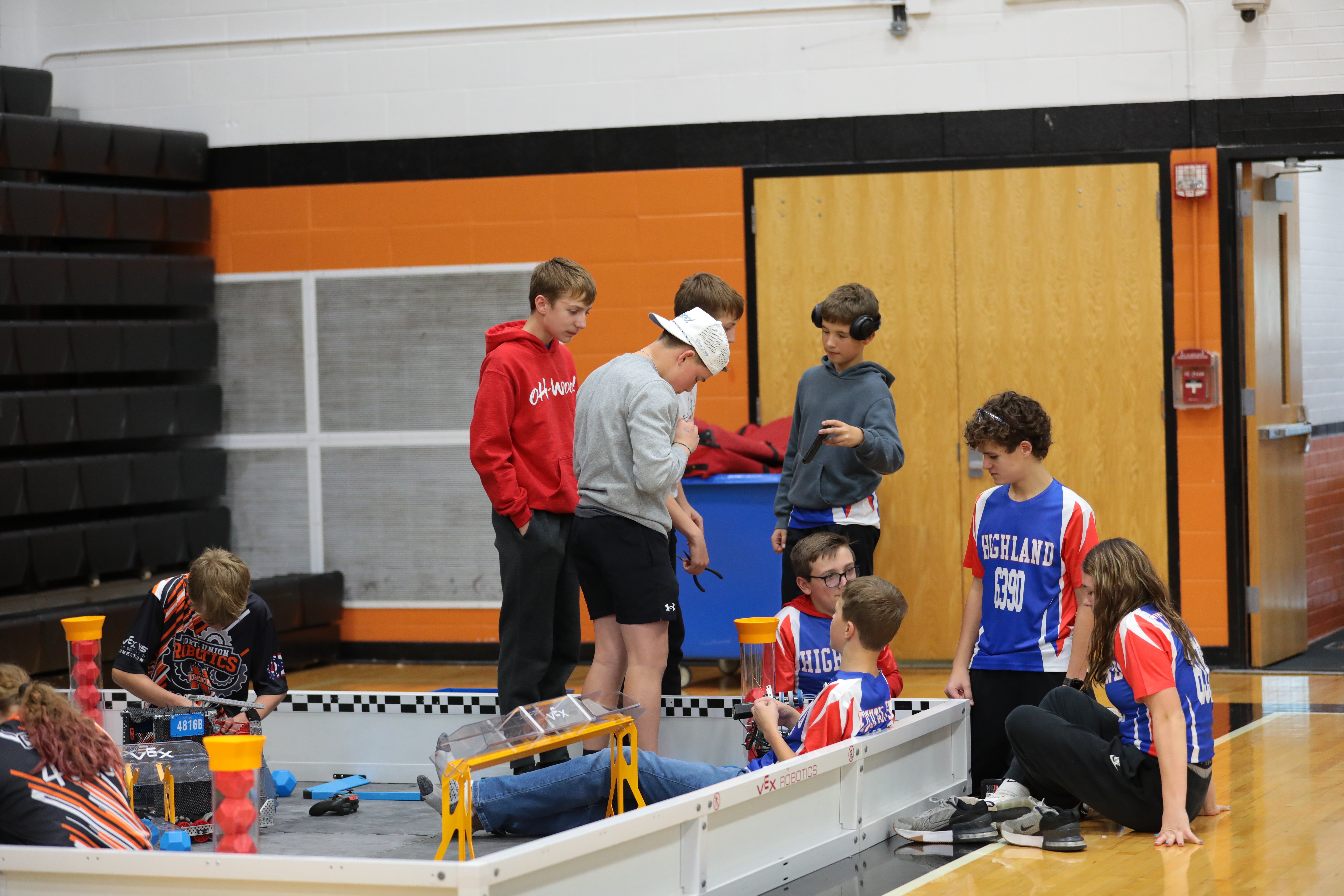 Teams of students sit and stand in and around a V5 competition field on the floor of a gym, preparing for matches at a league sesion.