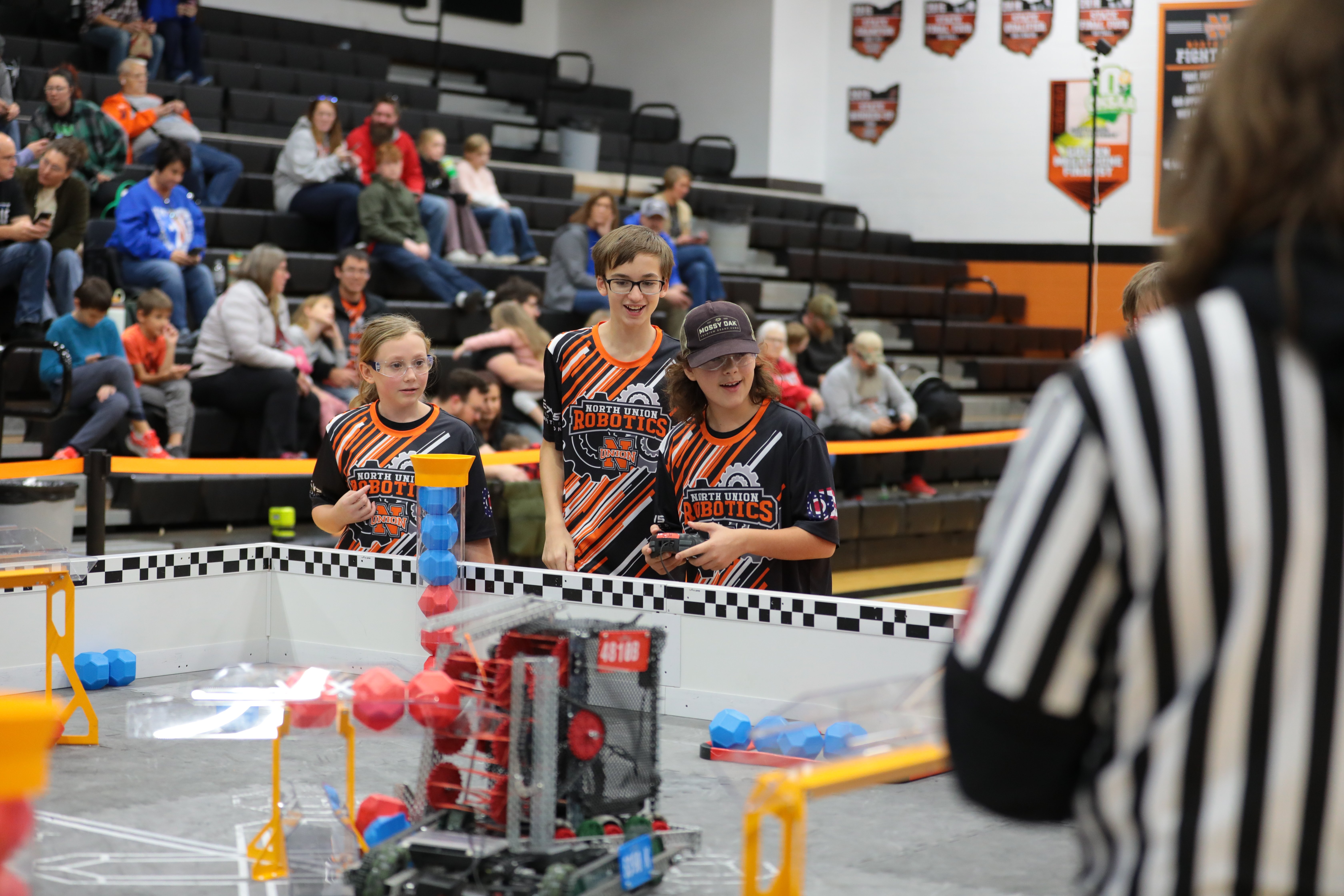 A team stands in the Driver's Station beside the V5 competition field, excited as they drive. In the foreground, a referee watches the match, and behind them spectators sit in bleachers.
