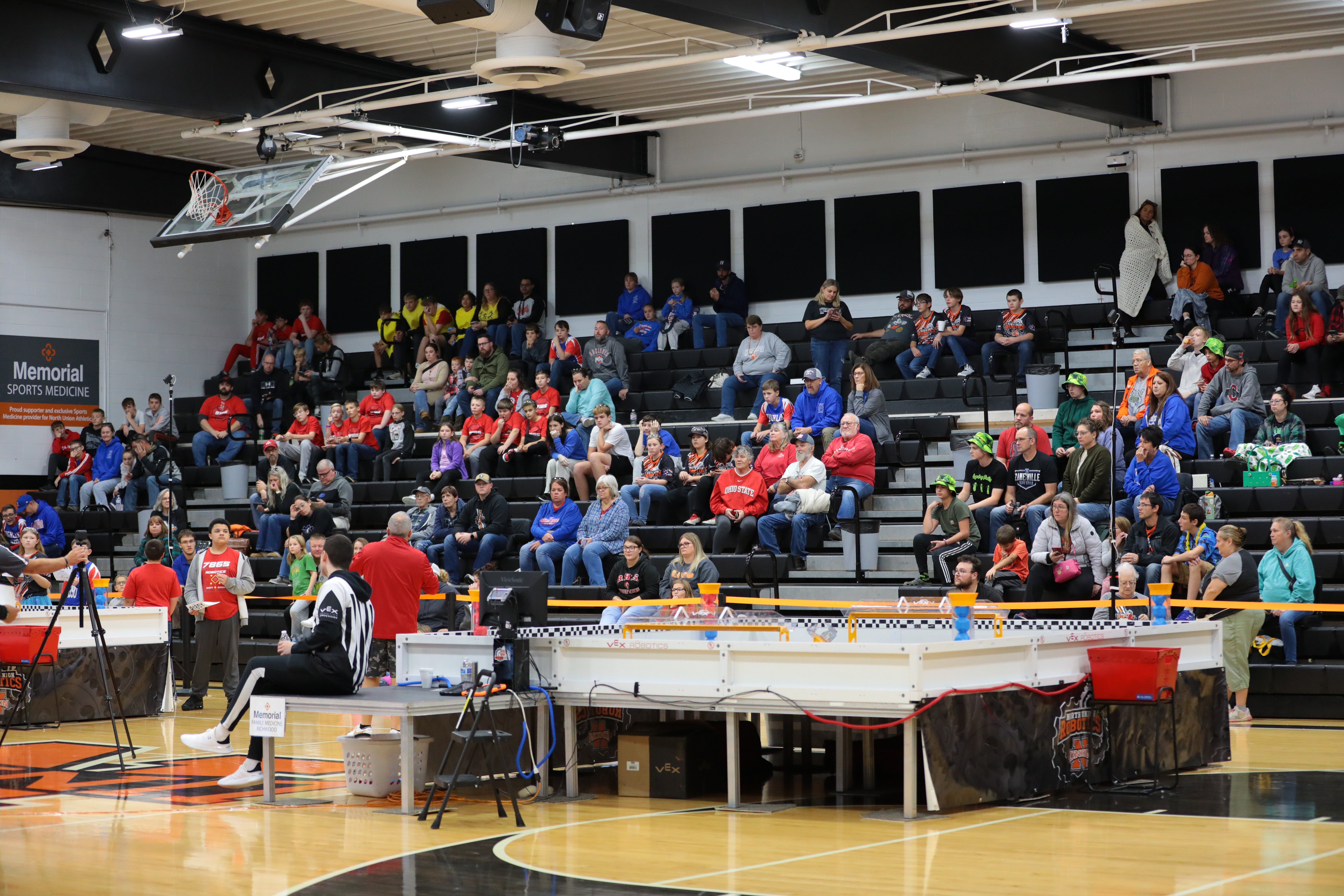 A wide view of a school gym set up for a VEX V5 competition. A competition field sits on tables on the court, with a camera tripod beside it, and spectators fill the bleachers behind it.