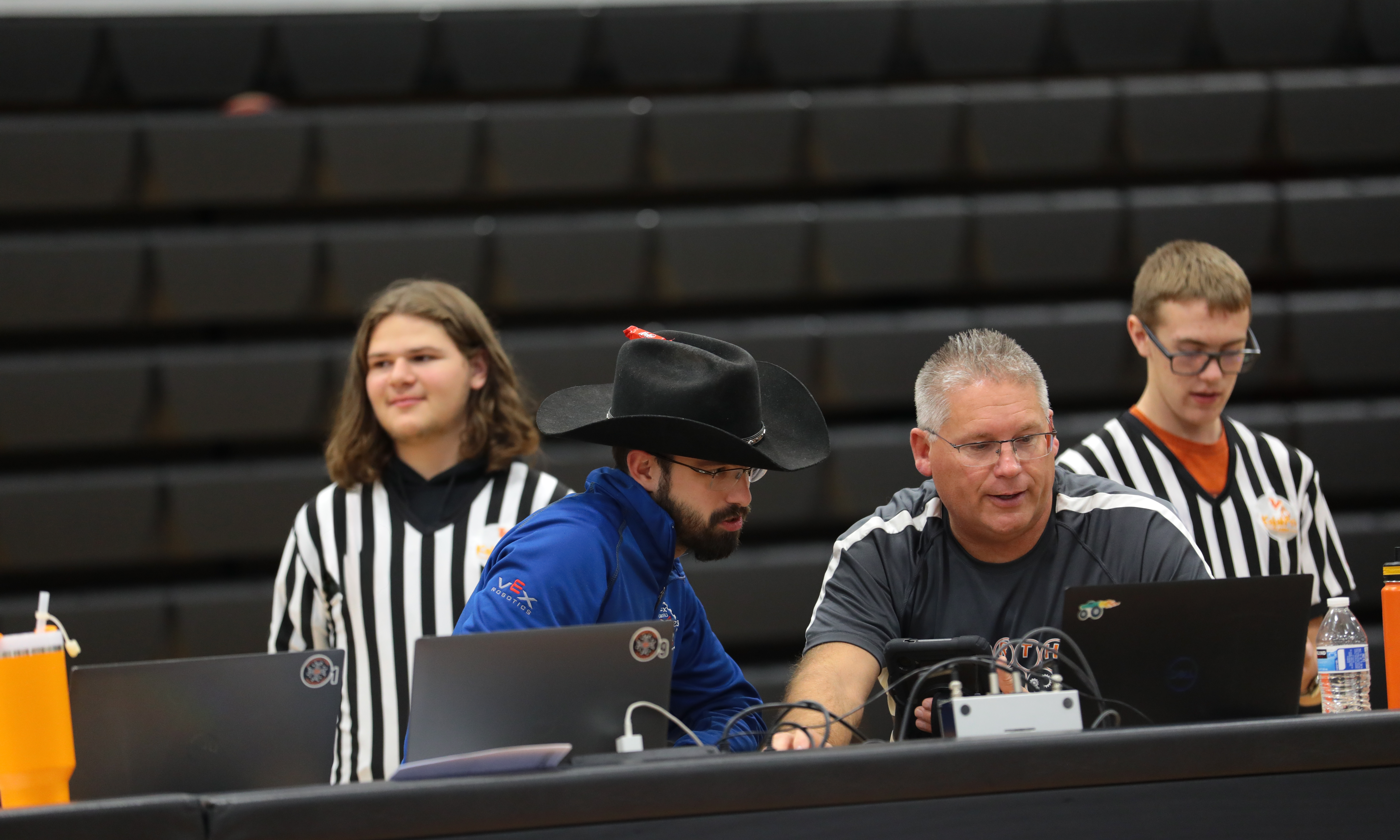 Two volunteers sit at a table with several laptops and devices, connecting a tablet to the computer so the referees standing behind them can use it to score a match.