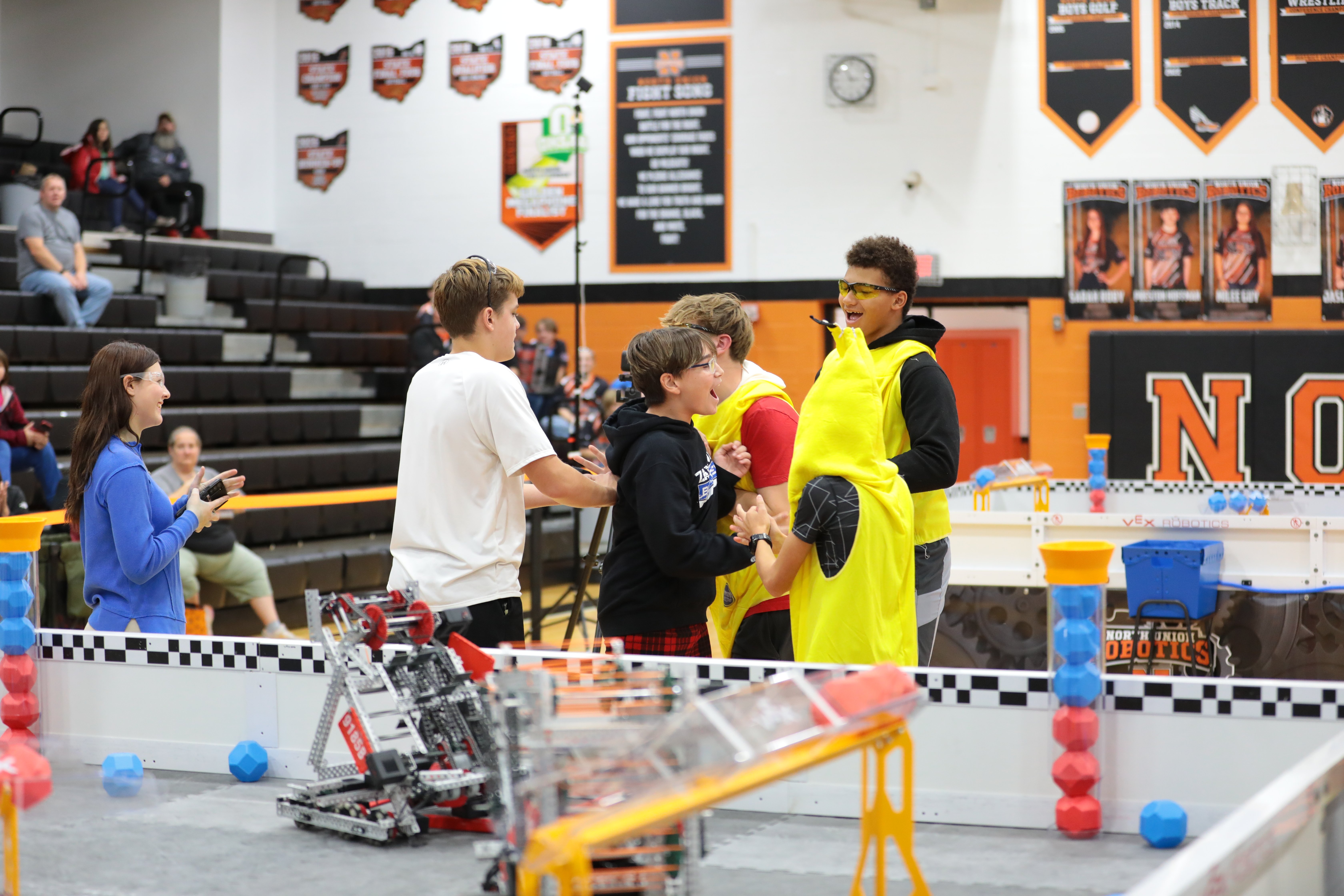 Teams celebrate after a match between two competition fields in a school gym, with spectators in the bleachers on the left and a volunteer clapping beside them.
