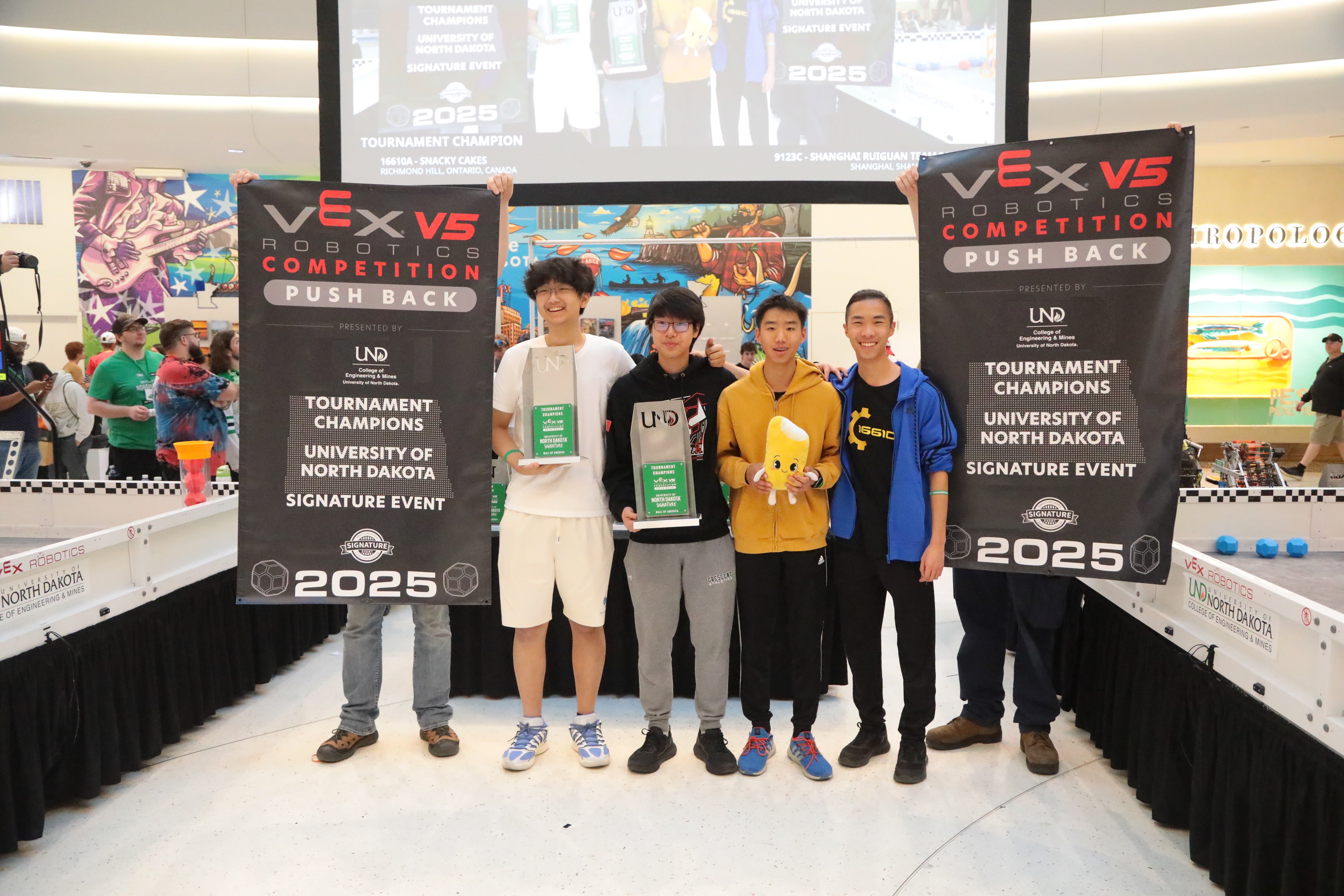 Four students representing two teams, stand smiling between two banners holding trophies. The banners indicate the teams are Tournament champions.