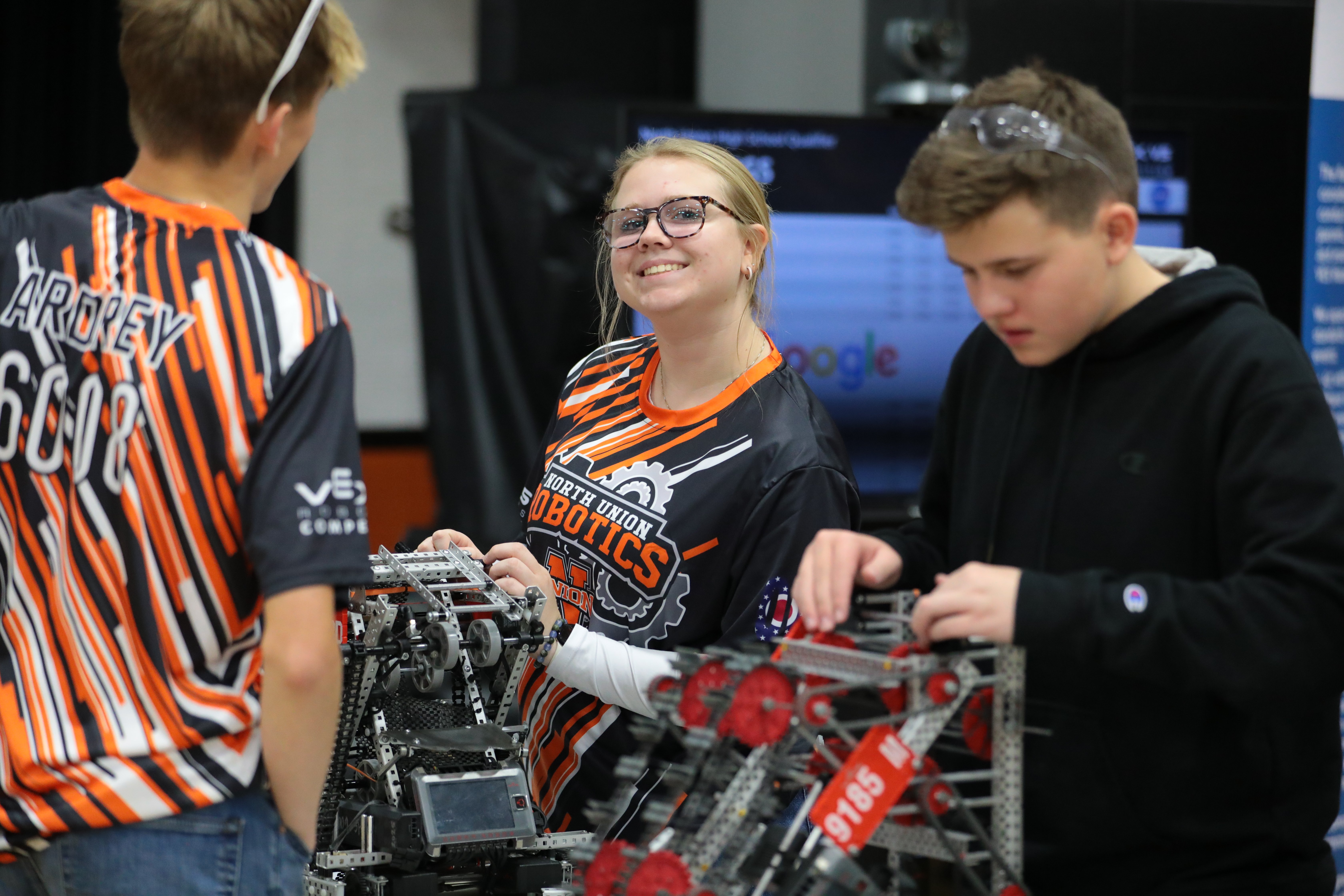 Three high school students gather around a VEX V5 robot at a pit table. A student in an orange-and-black team jersey smiles while holding the robot steady as a teammate in a black hoodie adjusts it. A display screen is visible behind them.