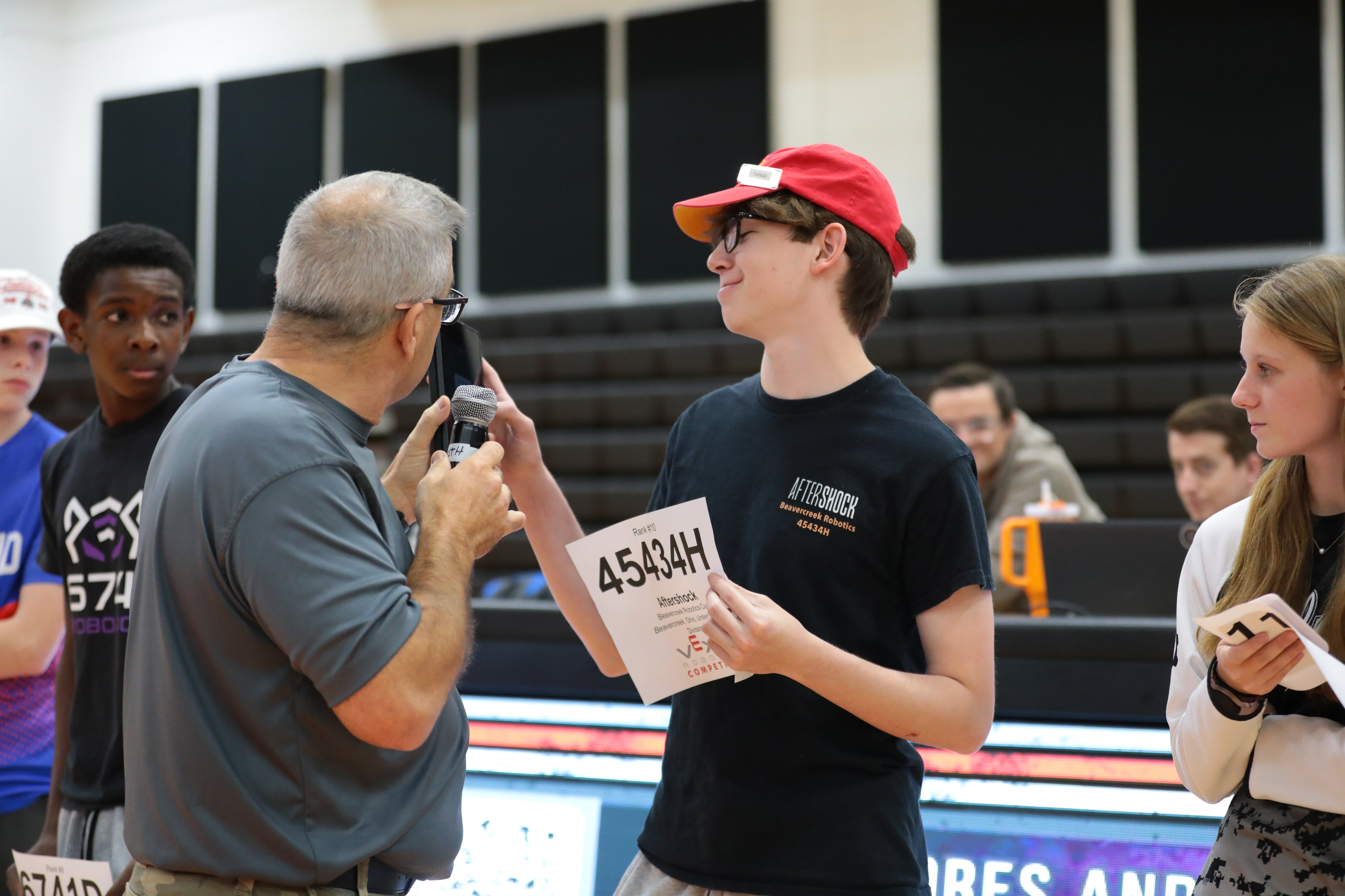 During Alliance Selection, the emcee holds a microphone toward a student in a red cap, who holds a printed sign showing his team number. Other team representatives with number signs stand nearby.