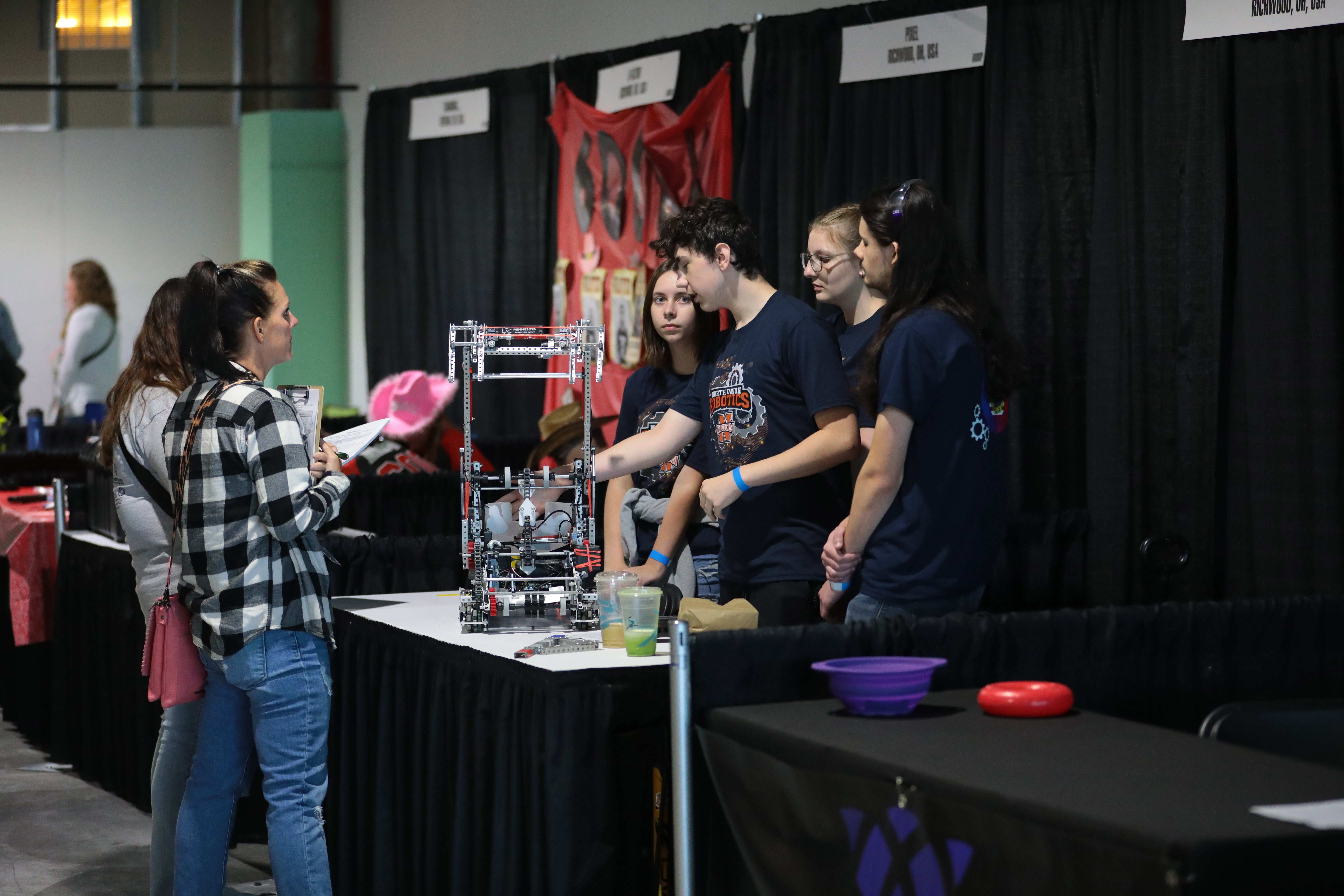 Two judges stand at a team's pit talking with a team. One student points at a feature of the robot while explaining, and the rest of the team looks on.