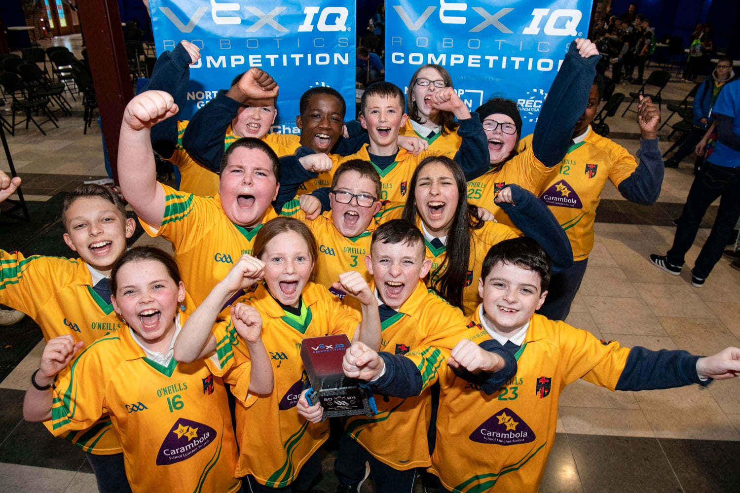 A team of boys and girls stand in front of two VEX IQ Robotics Competition banners cheers and celebrating together.