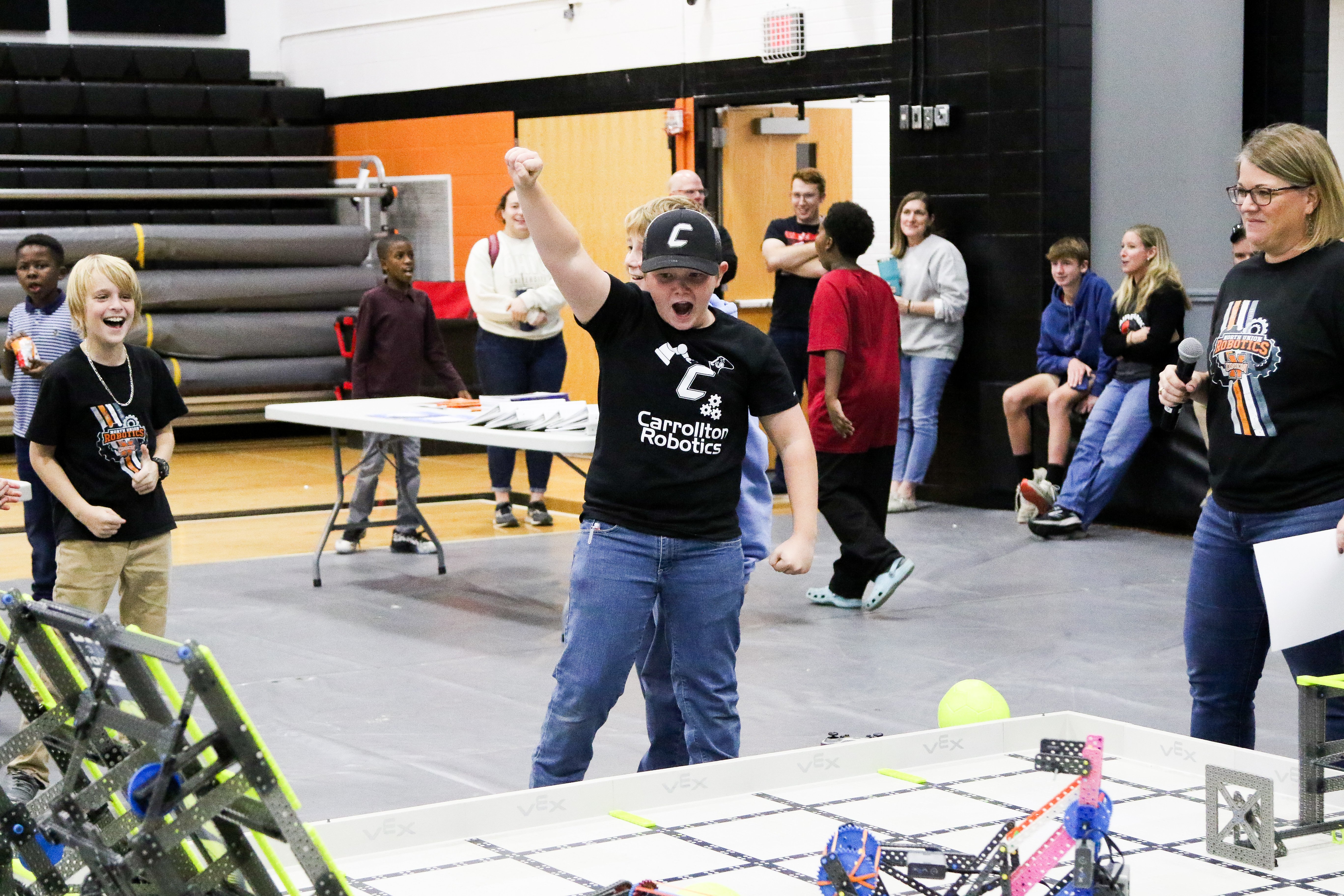 Students cheer beside an IQ competition field with a hand raised, as a smiling Emcee with a microphone looks on and spectators stand behind near queuing tables inside of a school gym.