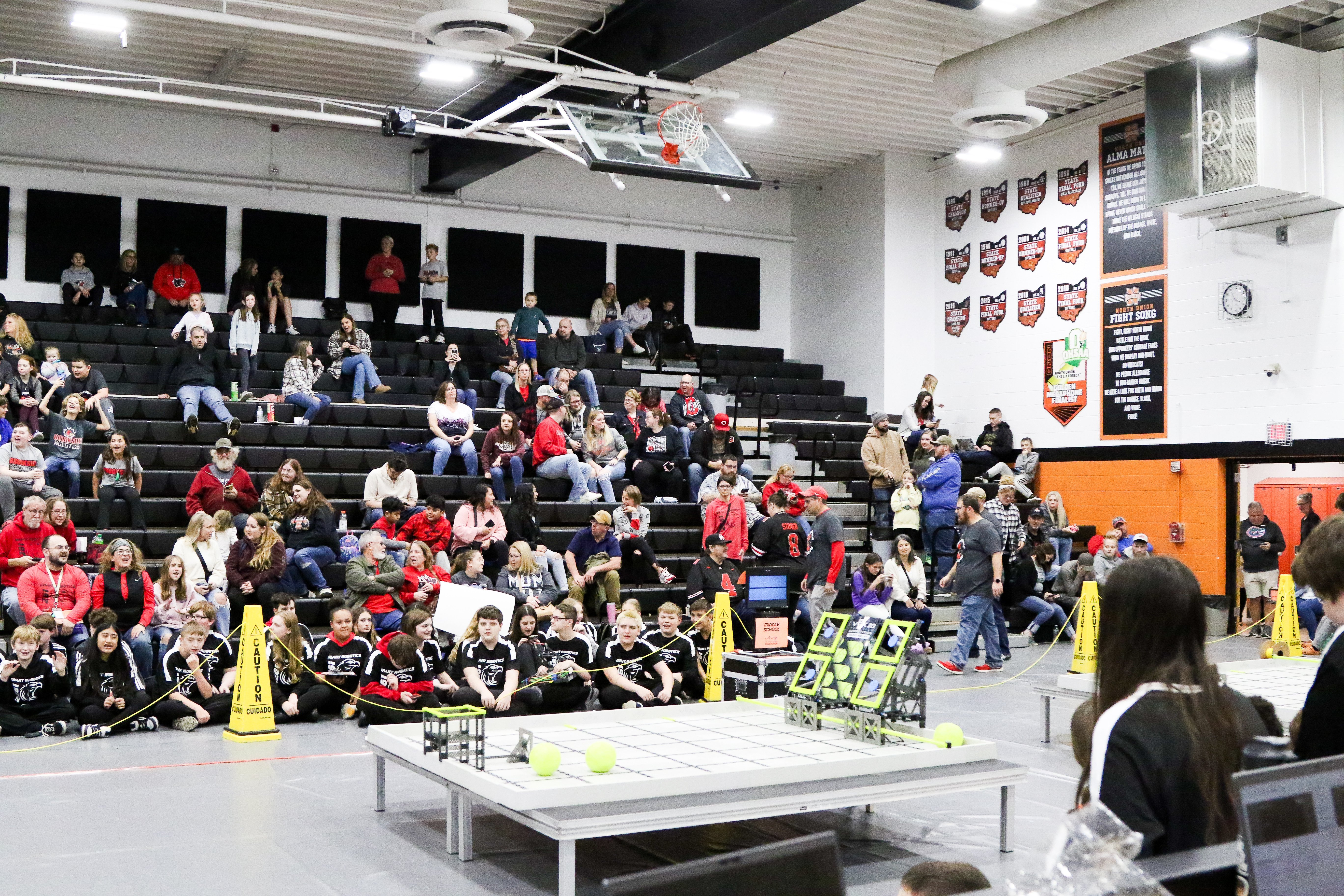 A wide view of a school gym set up for a VEX IQ competition. A competition field sits on a table in the foreground, marked off with stanchions and yellow caution cones. Teams sit on the floor in front of the bleachers, and spectators fill the seats above them.