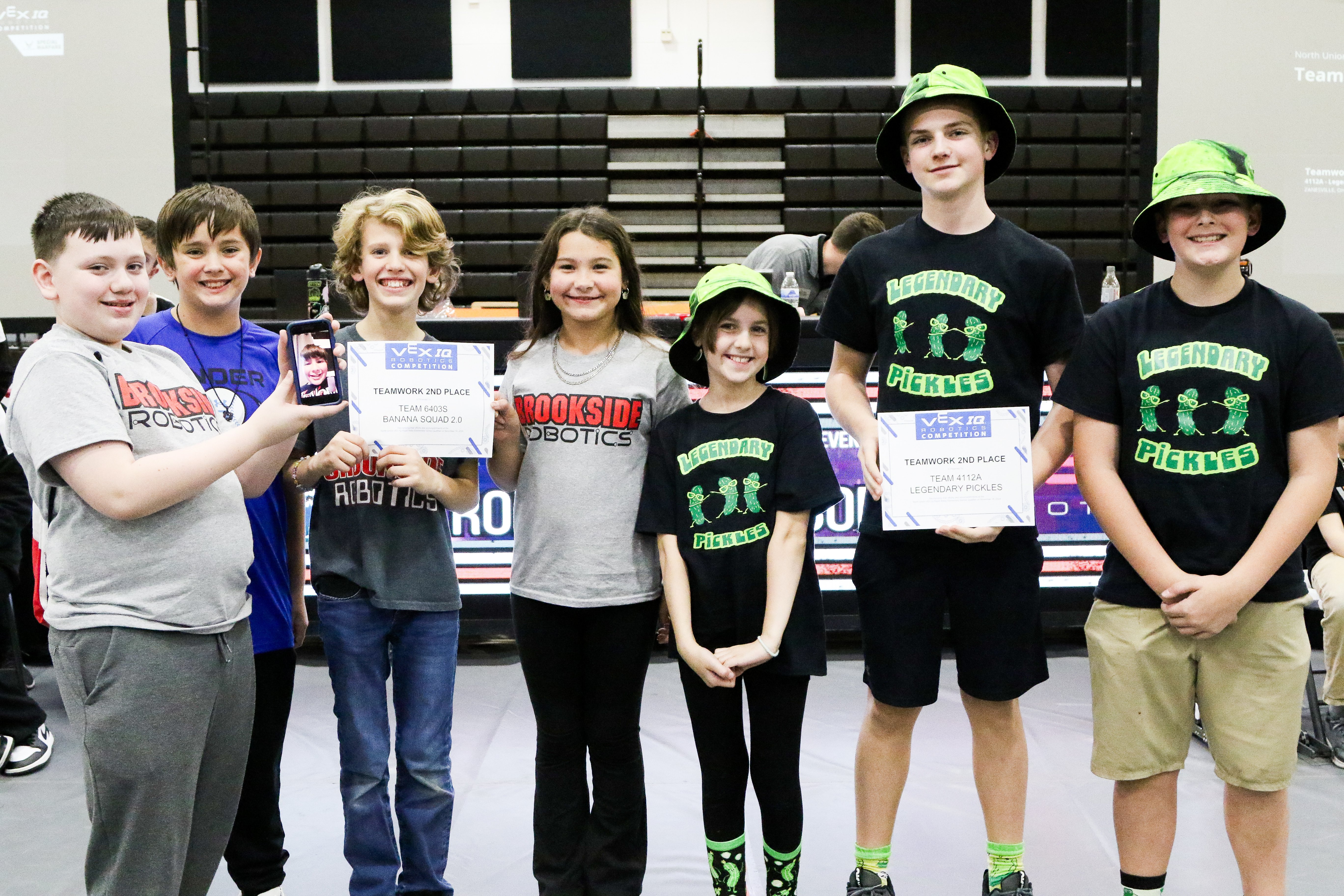 Students from two teams stand together after receiving their awards, two of them holding printed certificates. One team wears matching black shirts, several with green bucket hats.