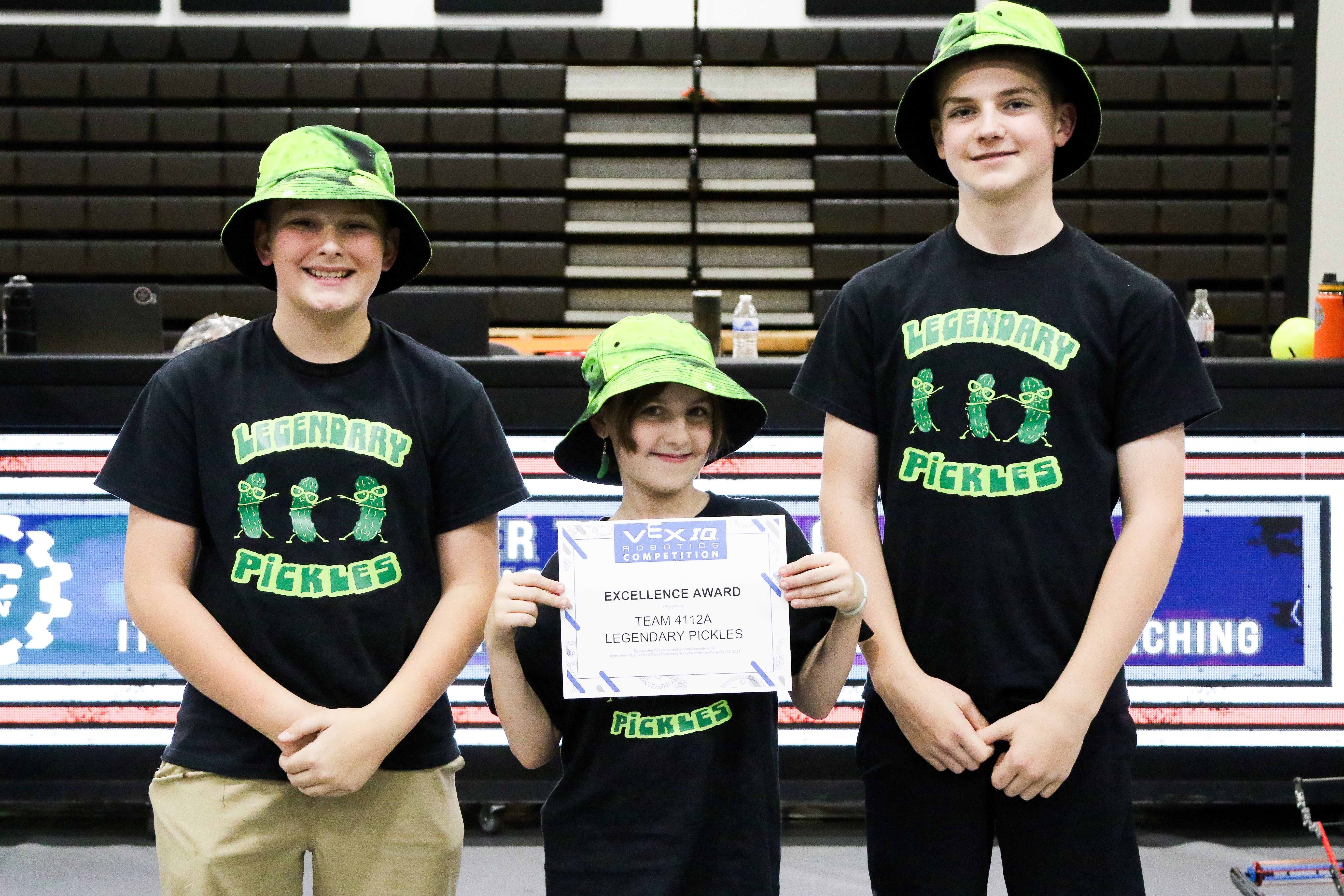 A team of three students pose for a photo after an Awards Ceremony. The girl in the center proudly holds their Excellence Award printed certificate.