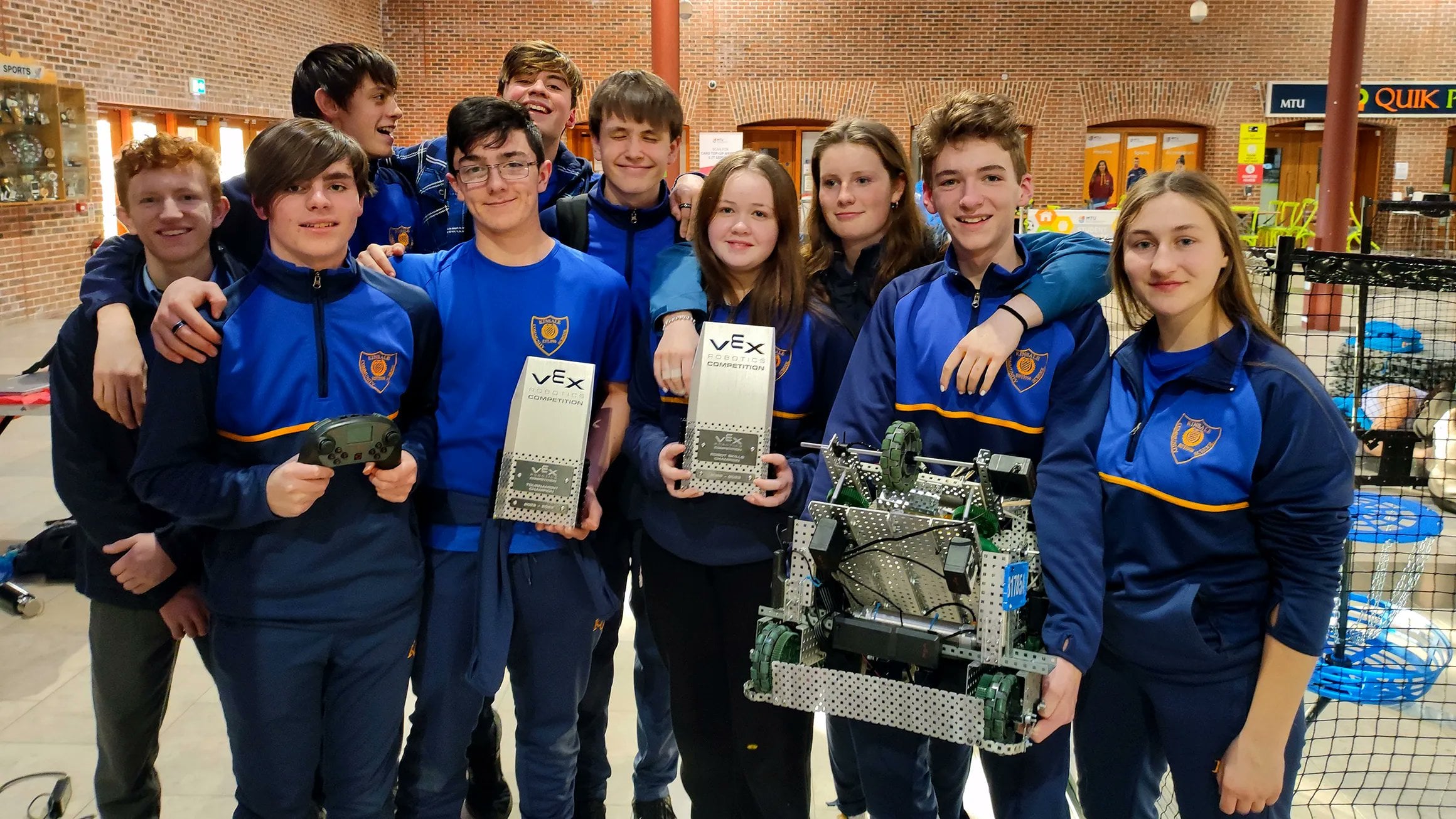 Two teams stand together, with arms around each other, holding their robots and each of their awards after a competition.