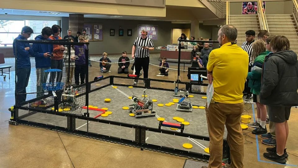 A V 5 match with two alliances on either side of the field. Also around the field are two scorekeeper referees in black and white striped shirts and a head referee in a yellow shirt.