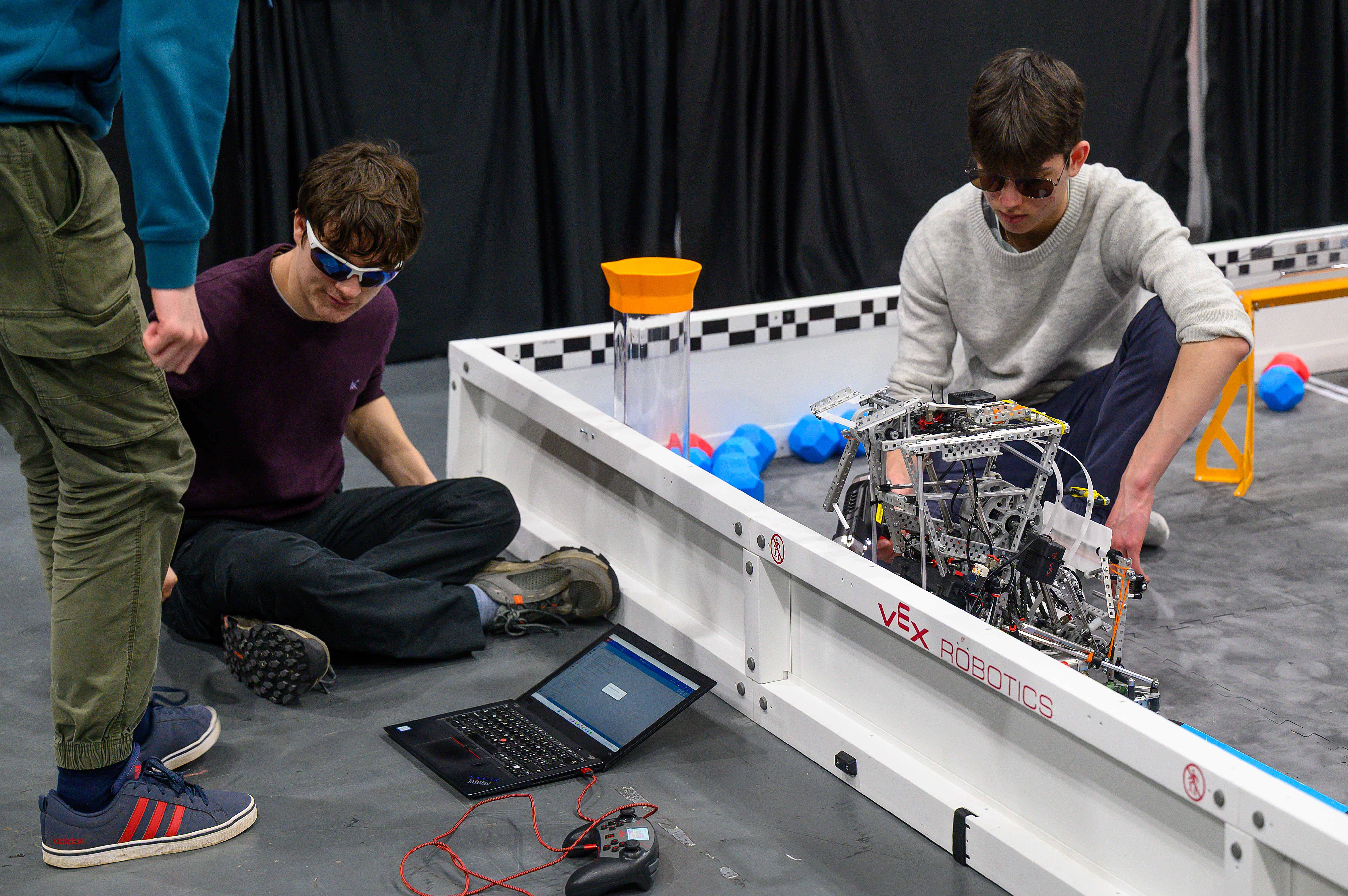Three team members work at a Field with one of their robos. One is on the field, adjusting the robot, a second is sitting on the floor beside the field with a laptop open and controller connected, while the third looks on beside them.