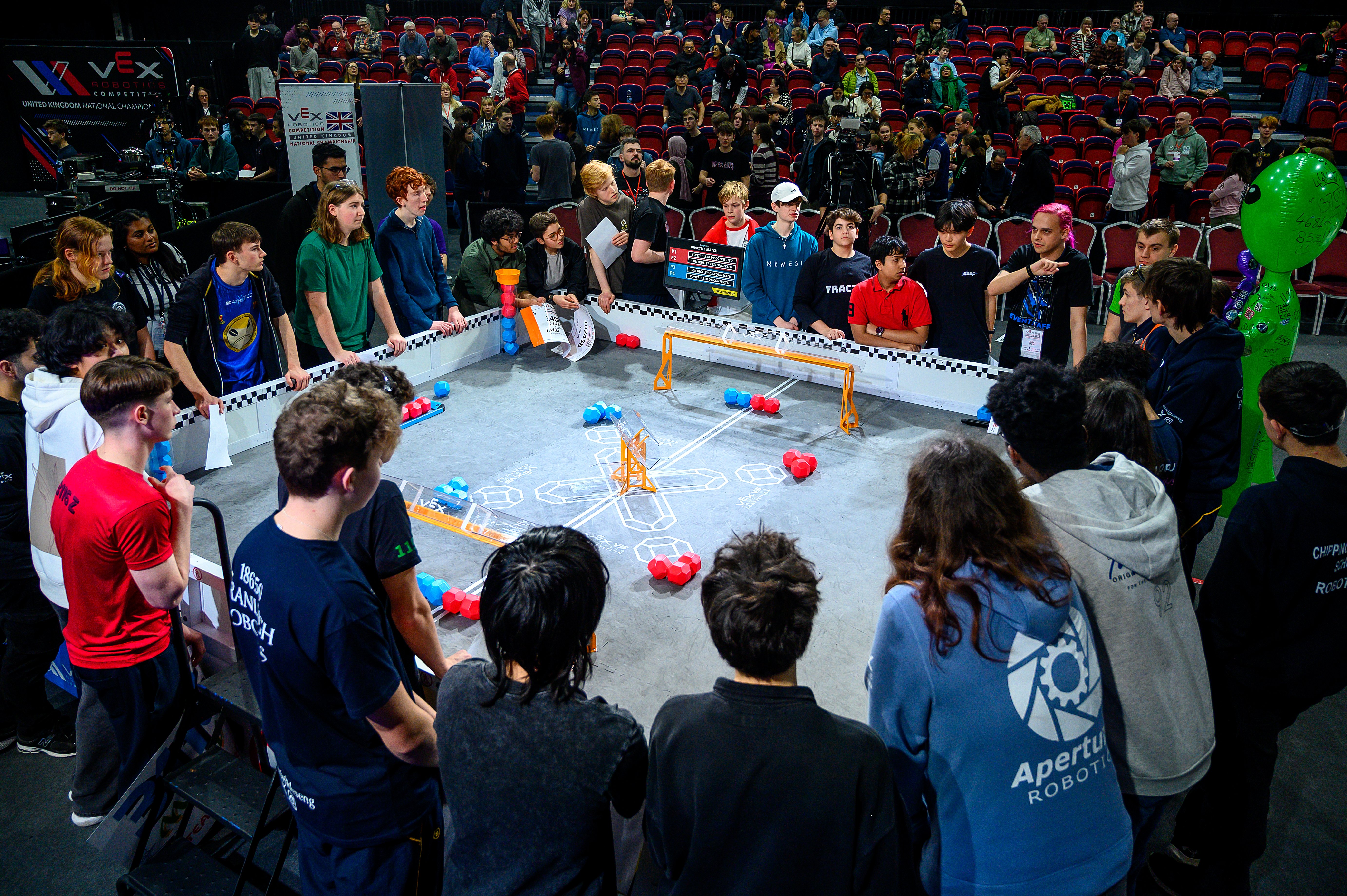 Team representatives stand around a competition field, as spectators look on for an Event Meeting. The Head Referee is speaking, and everyone else is listening.