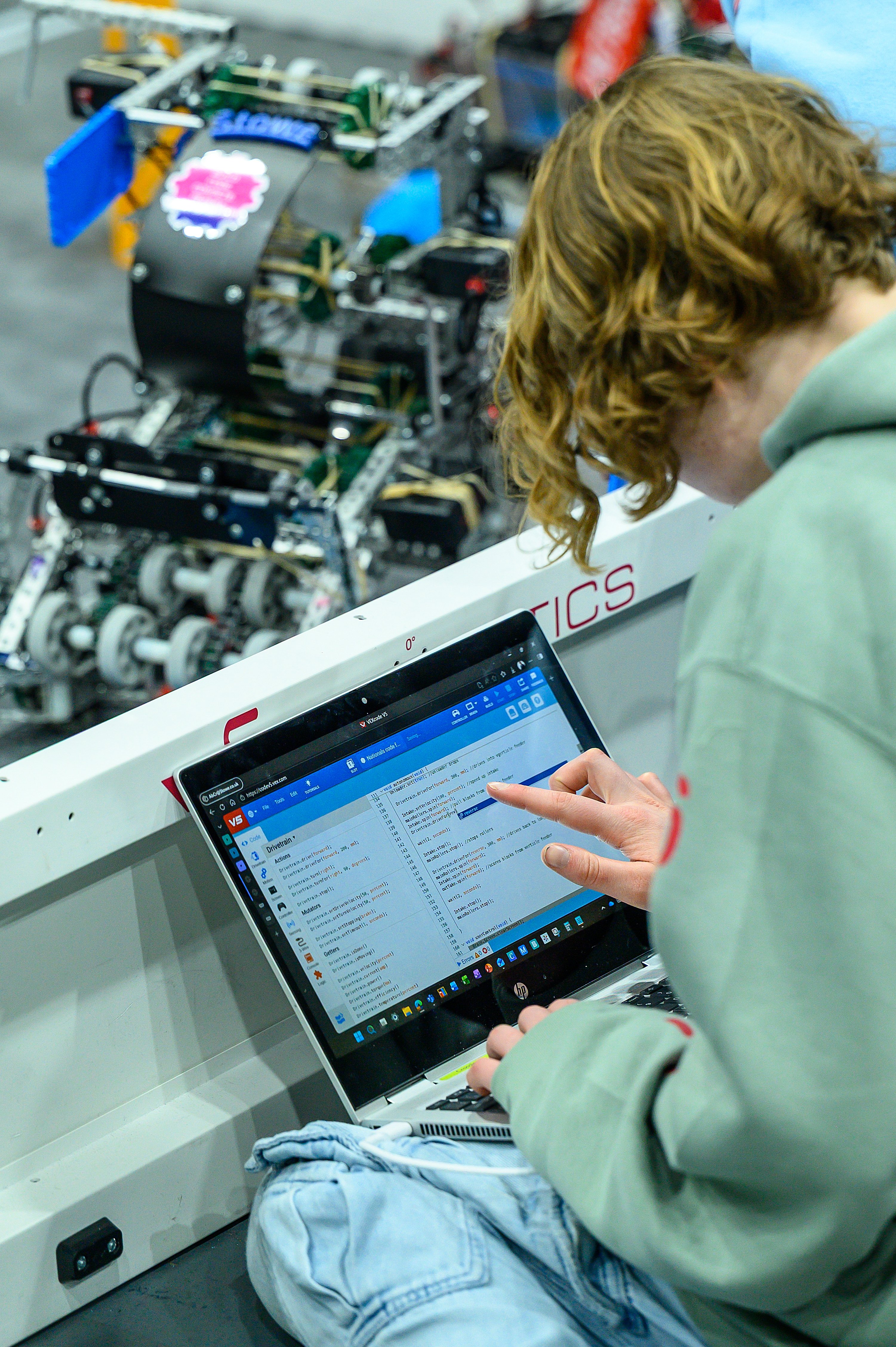 A student sits beside a Skills Field with the robot on the Field, reading at a VEXcode V5 project on a laptop.