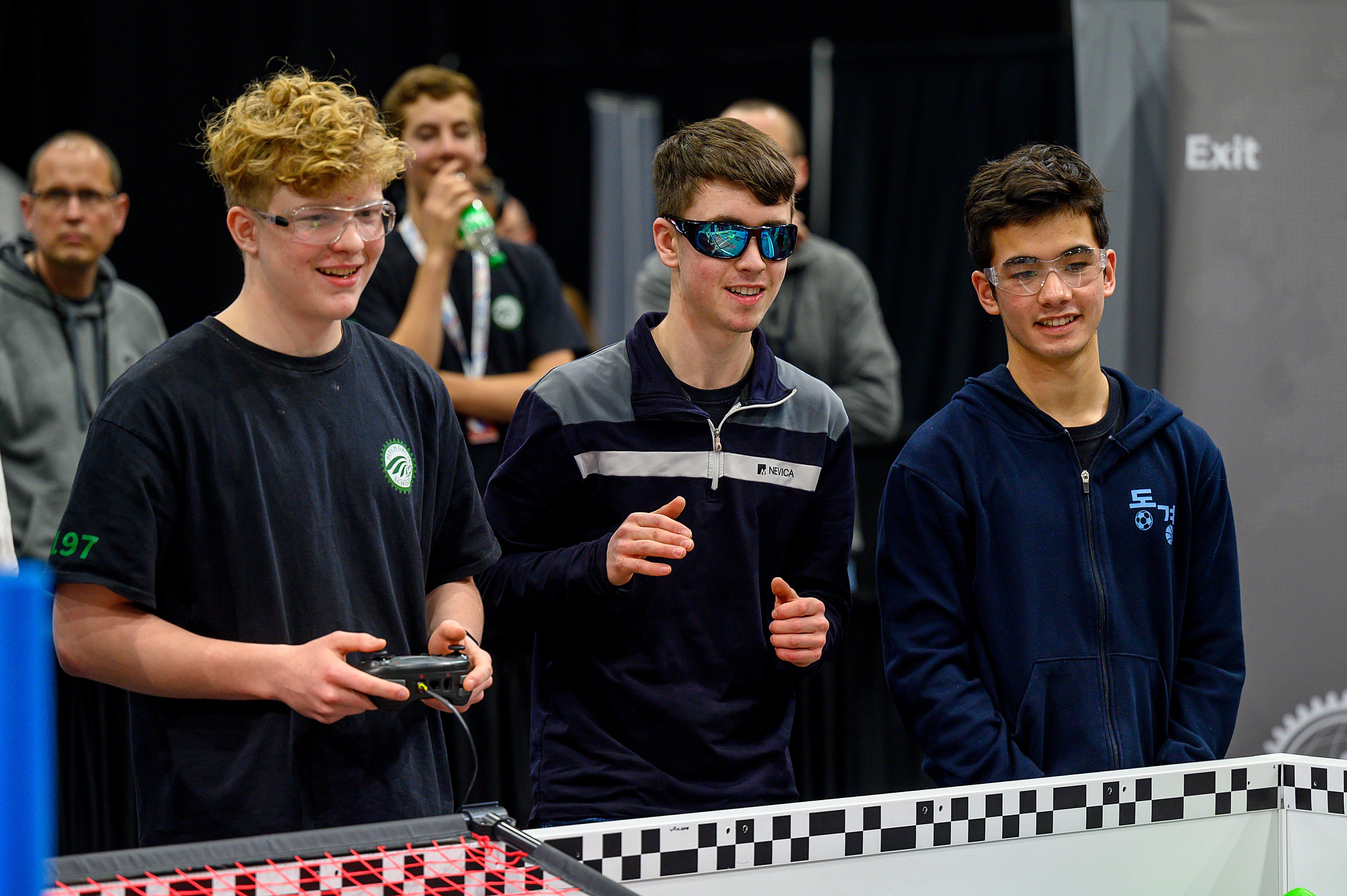 Three VEX U students stand alongside a Field with one holding a controller, and all three smiling and watching the match unfold.