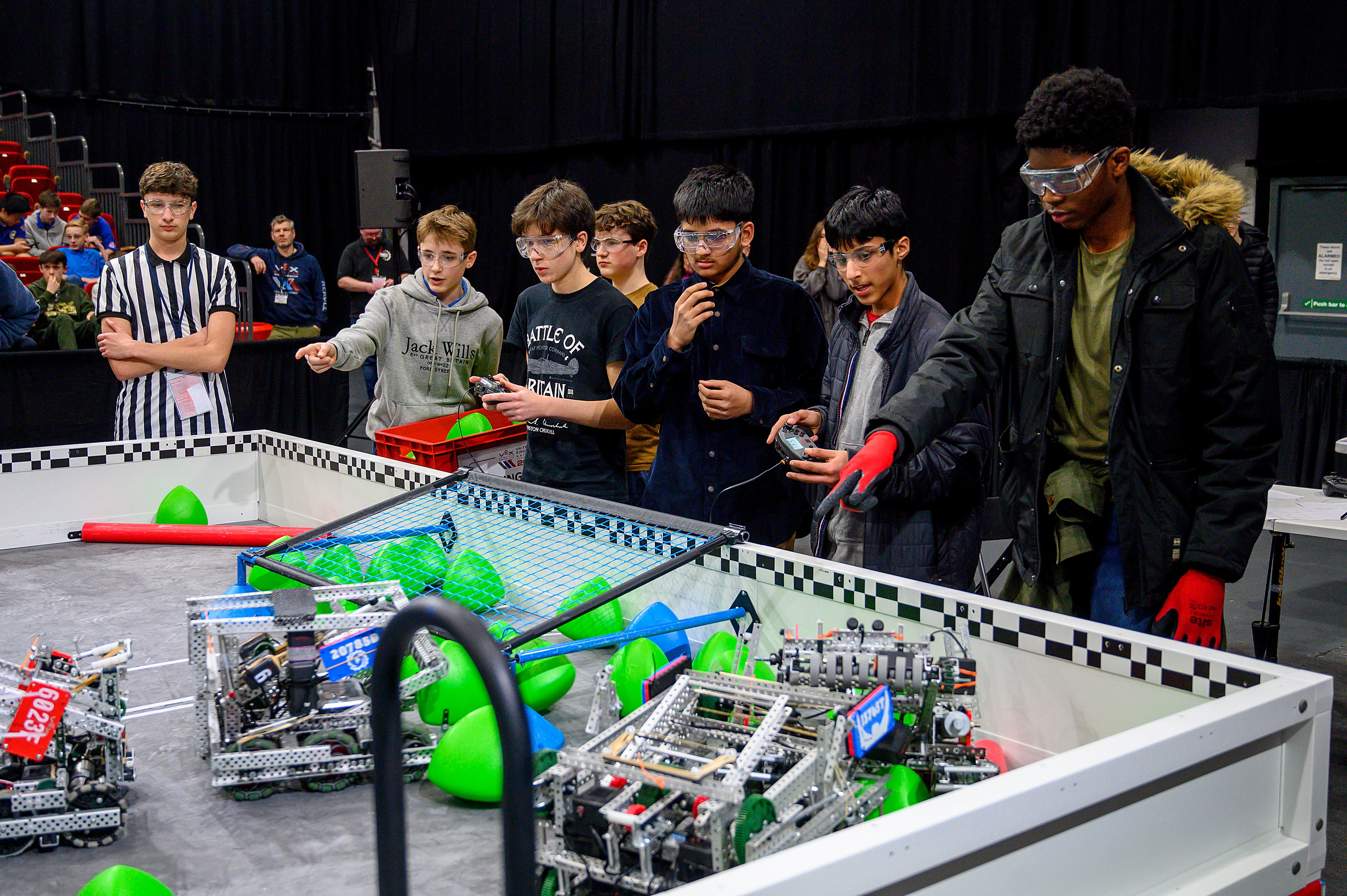 A V5 match in progress. All four robots can be seen in close proximity on the field. A referee looks on at the interactions from the background.
