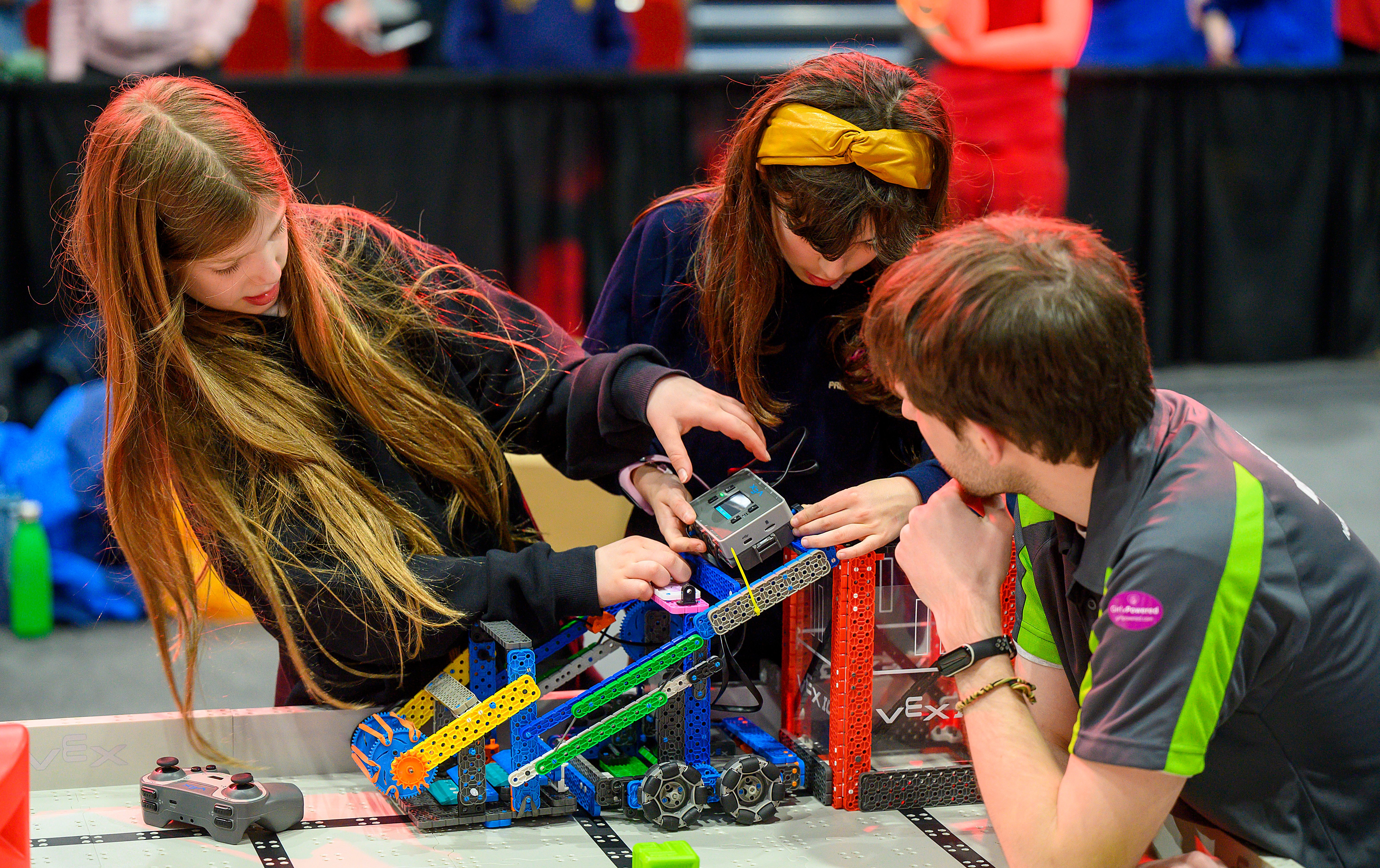 Two female Drive Team Members reset a robot on the field while the Referee looks on closely.