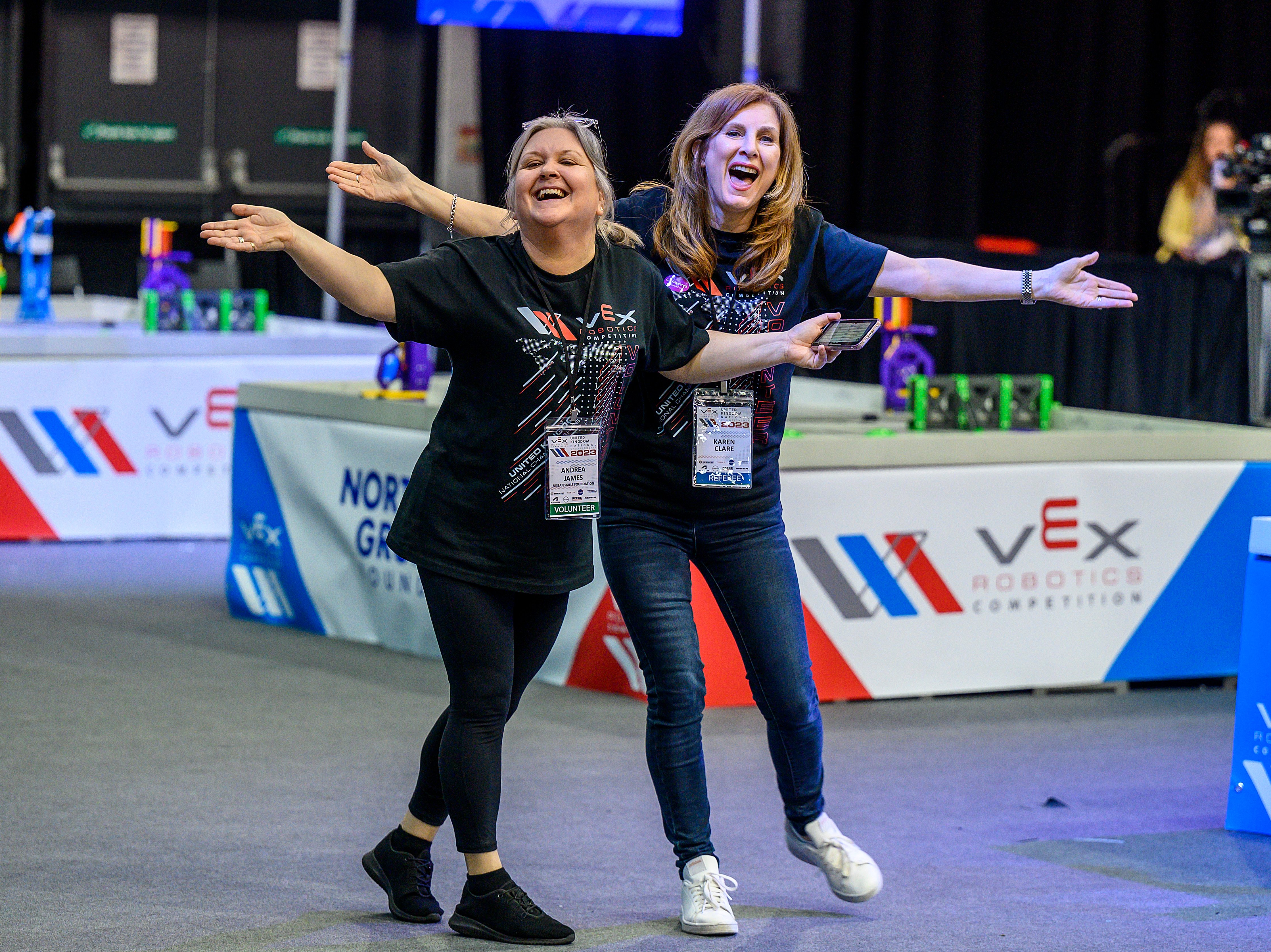 Two volunteers wearing lanyards and event badges laugh and throw their arms out wide in front of a competition field.