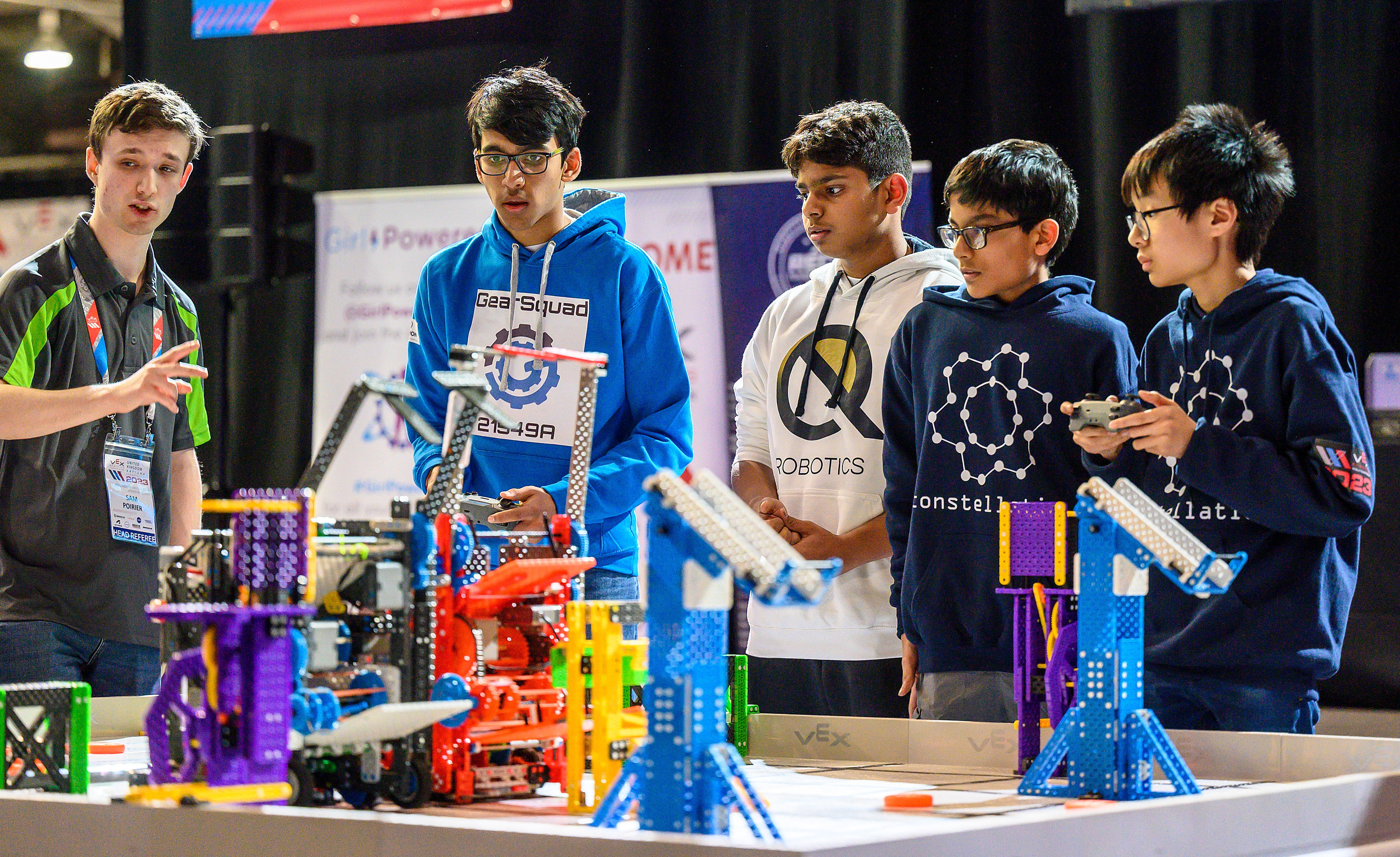 A Head Referee officiating a match and doing a count as two teams drive their robots on the field.
