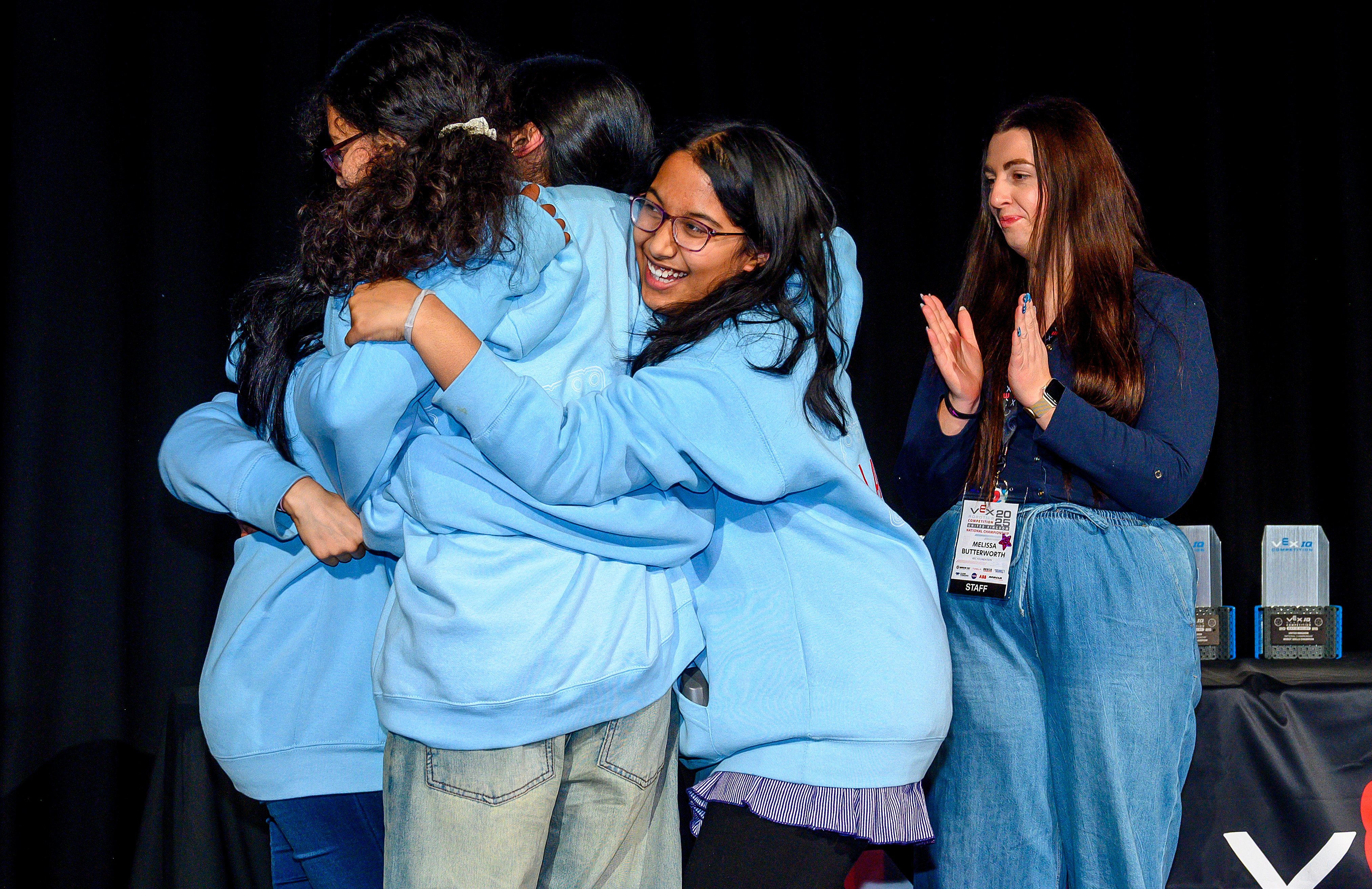 A team of girls in matching shirts jumps and hugs each other on stage, while a presenter claps in front of a table full of awards.