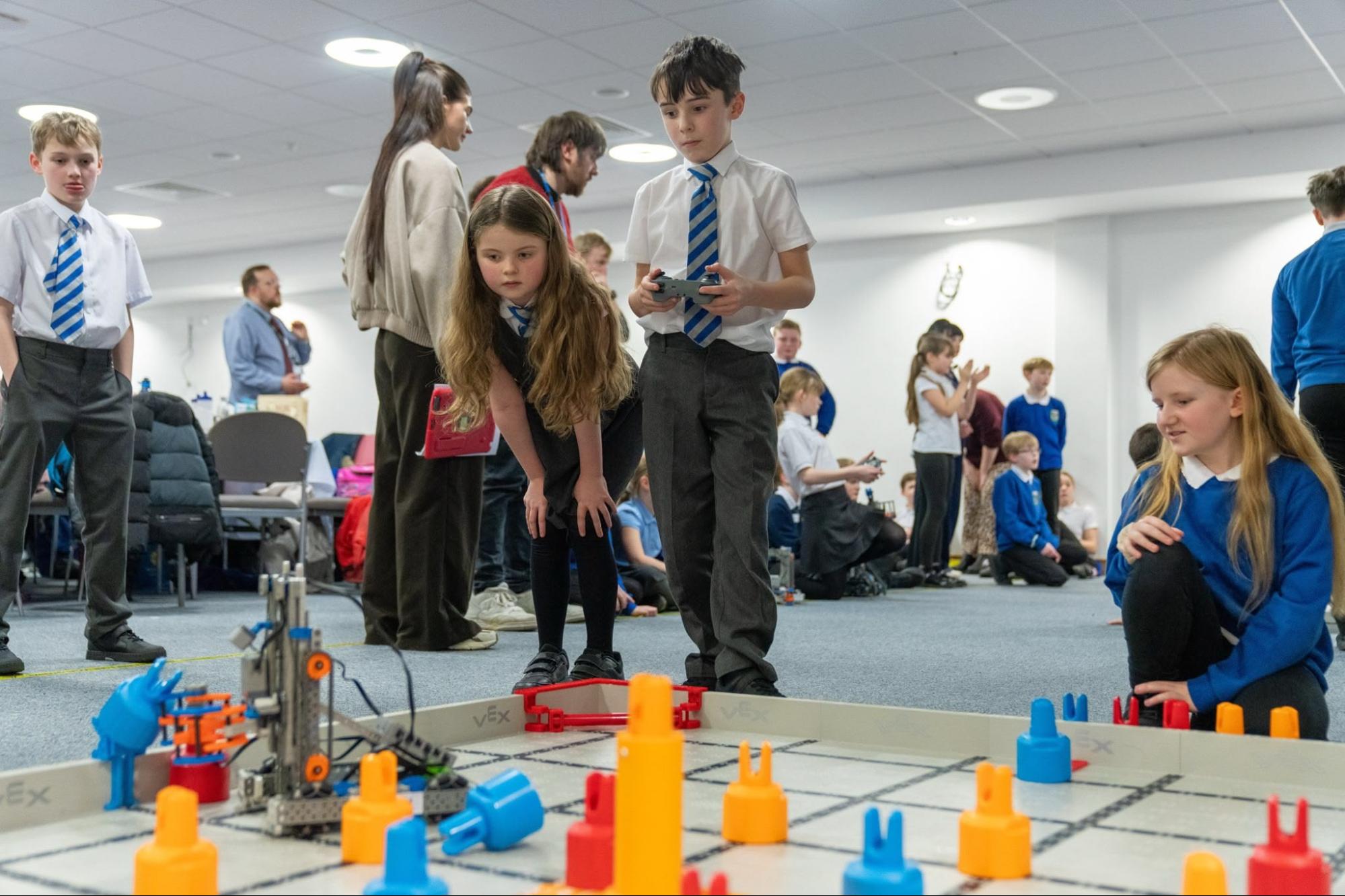 Students in school uniforms gather around a VEX IQ field set up on the carpet of a bright room, one driving while the others watch.
