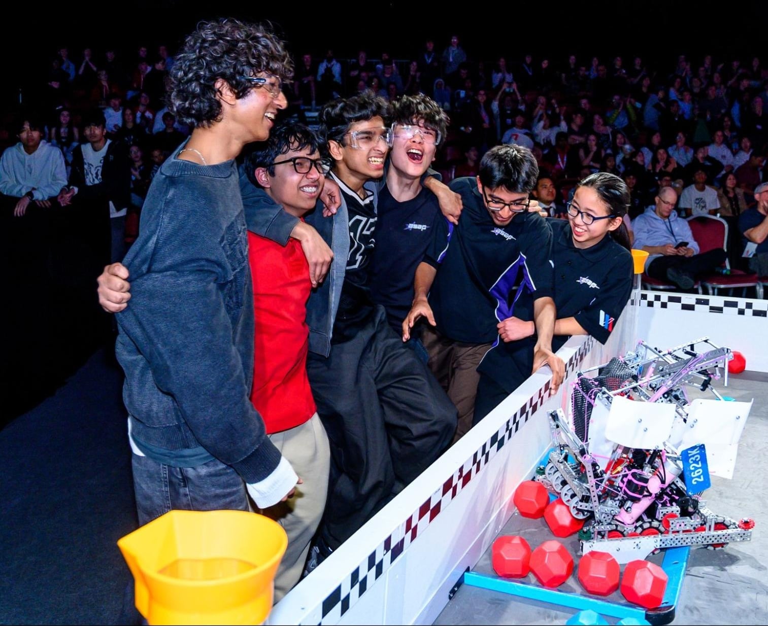 Six high school students link arms and celebrate beside their robot at the edge of a competition field, with a full audience behind them.
