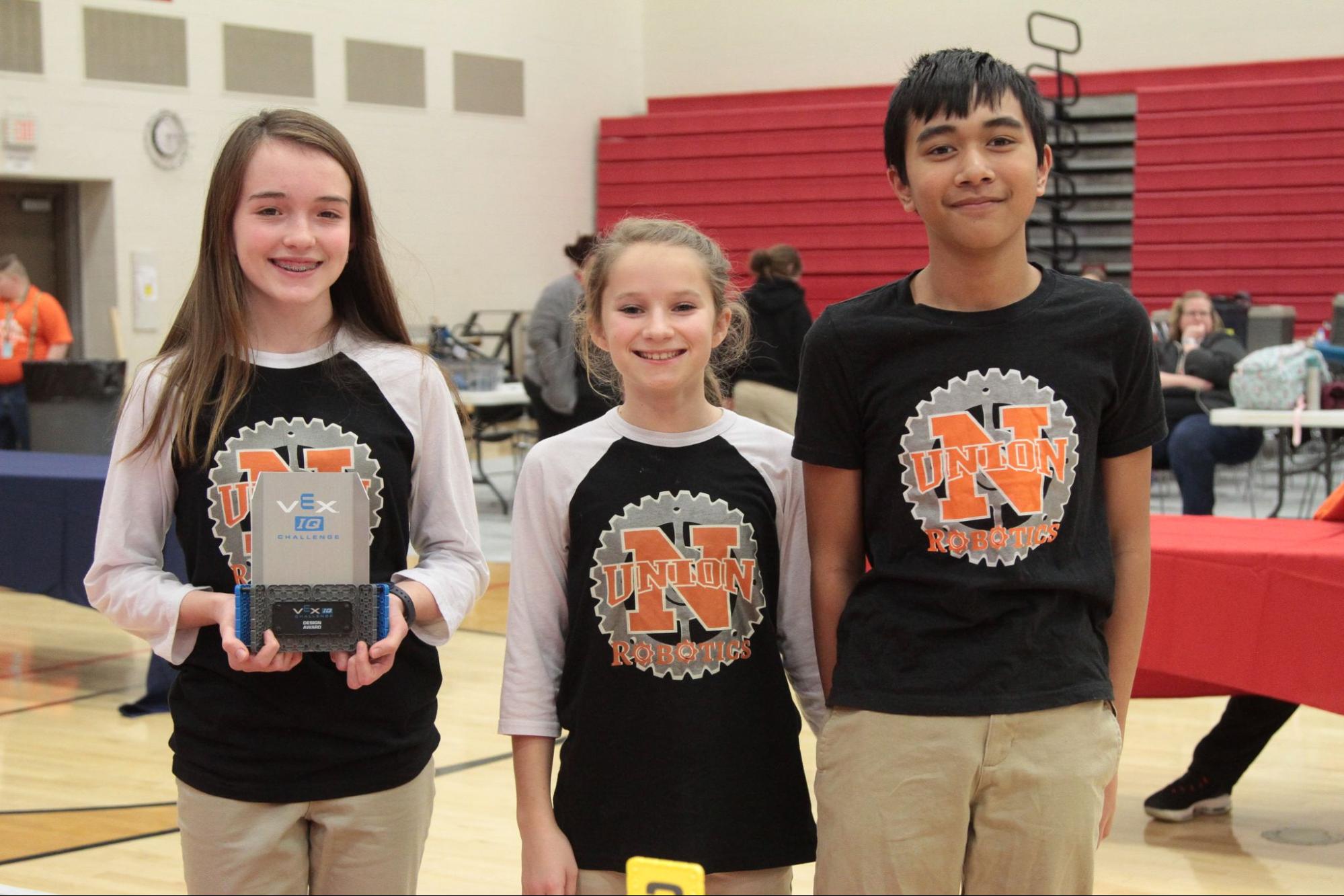 Three VEX IQ team members stand together in a school gym, the student in front holding up their team's award trophy.