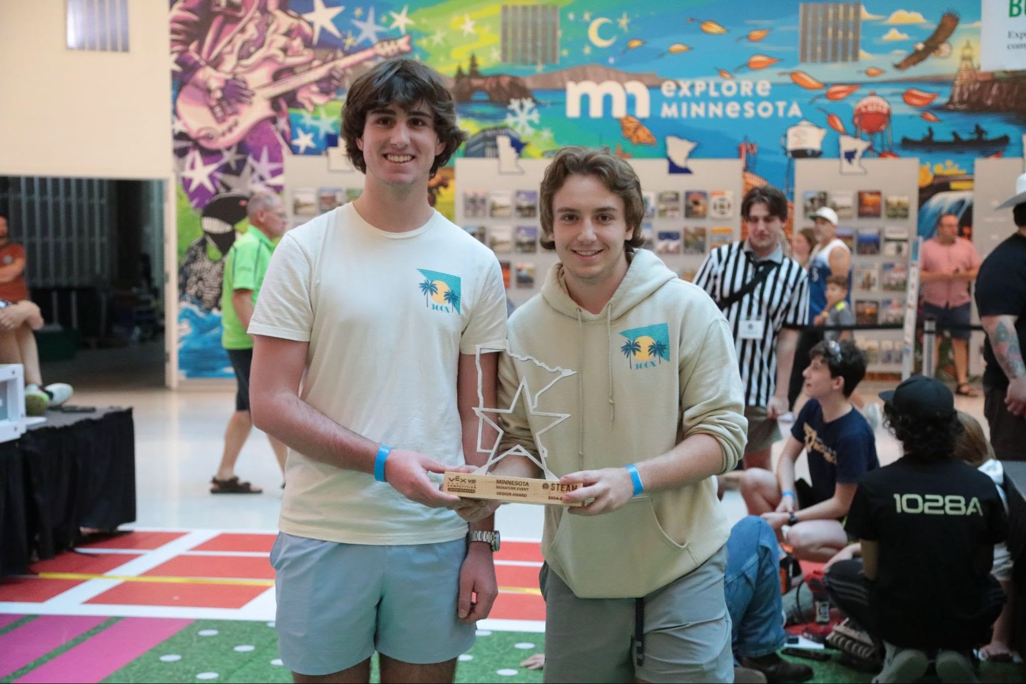Two students on a team stand in the middle of an event holding their Design Award trophy and smiling.