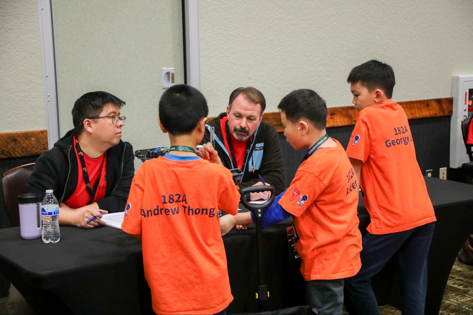 A team of three students stand at a Judge's table talking with two judges. One points to something on the robot as he explains.