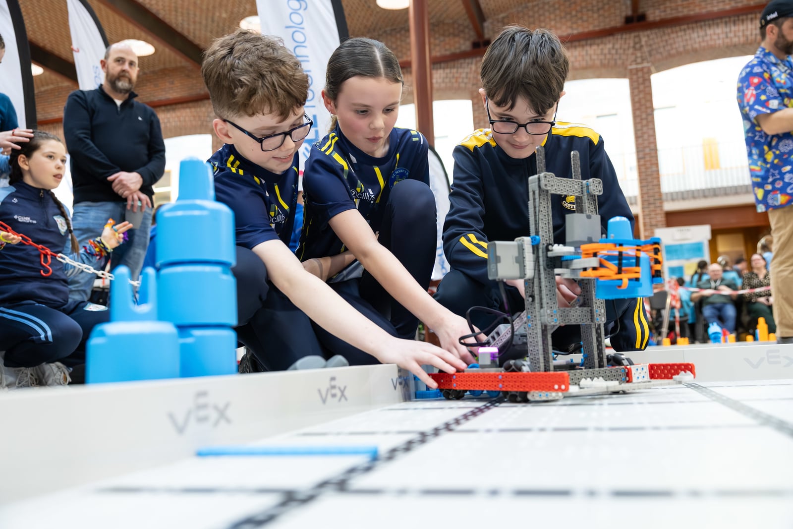 Three students crouch at a VEX IQ field to position their robot while adults stand back and watch.