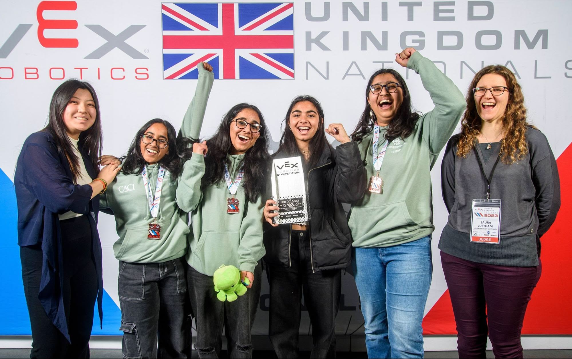 A team of girls stands in front of the United Kingdom Nationals VEX Robotics tournament sign cheering while holding their award, alongside the judges.