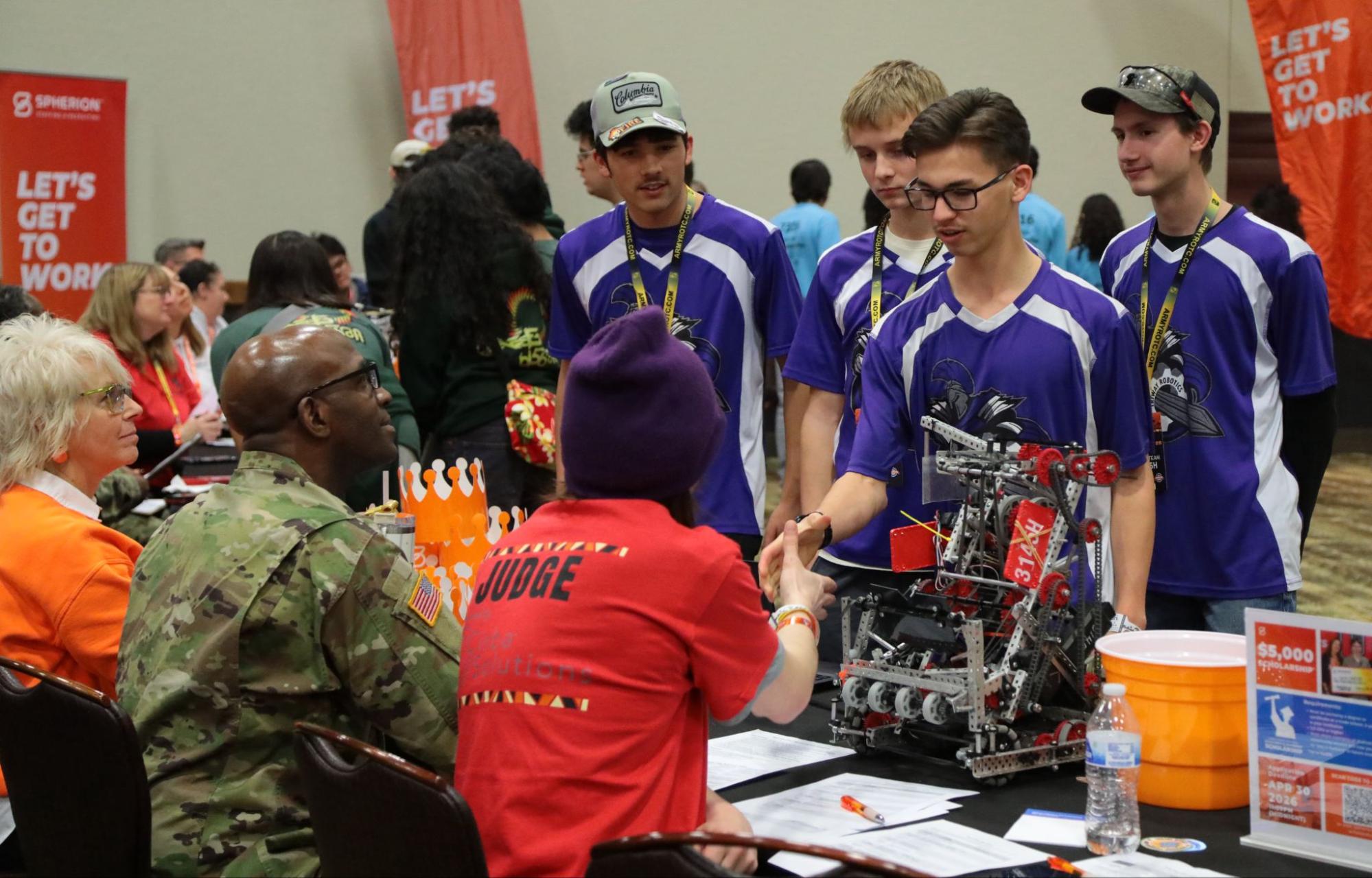 A team of older students greets a judging team at their table, shaking hands with the judge as they place the robot on the table at the start of the interview.
