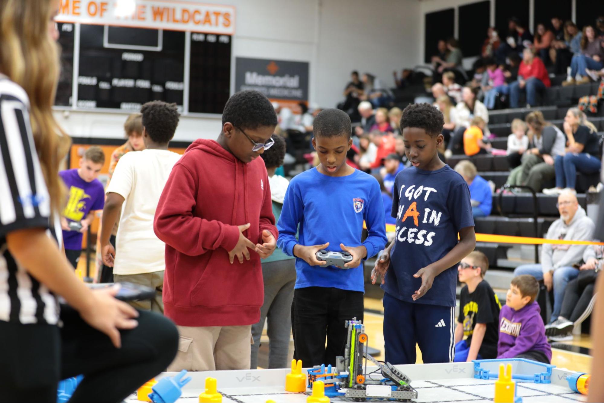 Three students stand at a VEX IQ field in a school gym, one holding a controller, with a referee beside them and spectators in the bleachers.