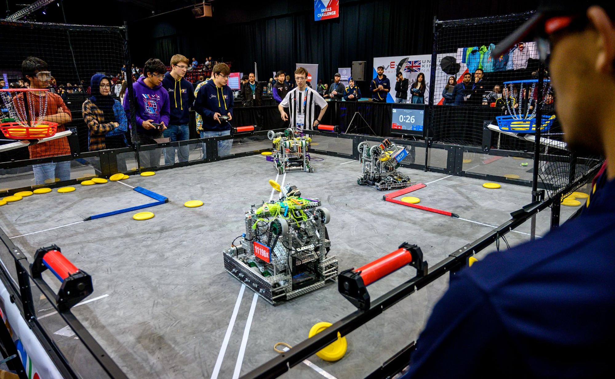 Students in safety glasses line one side of a VEX V5 field during a match, two of them driving with controllers while a referee watches from across the field.