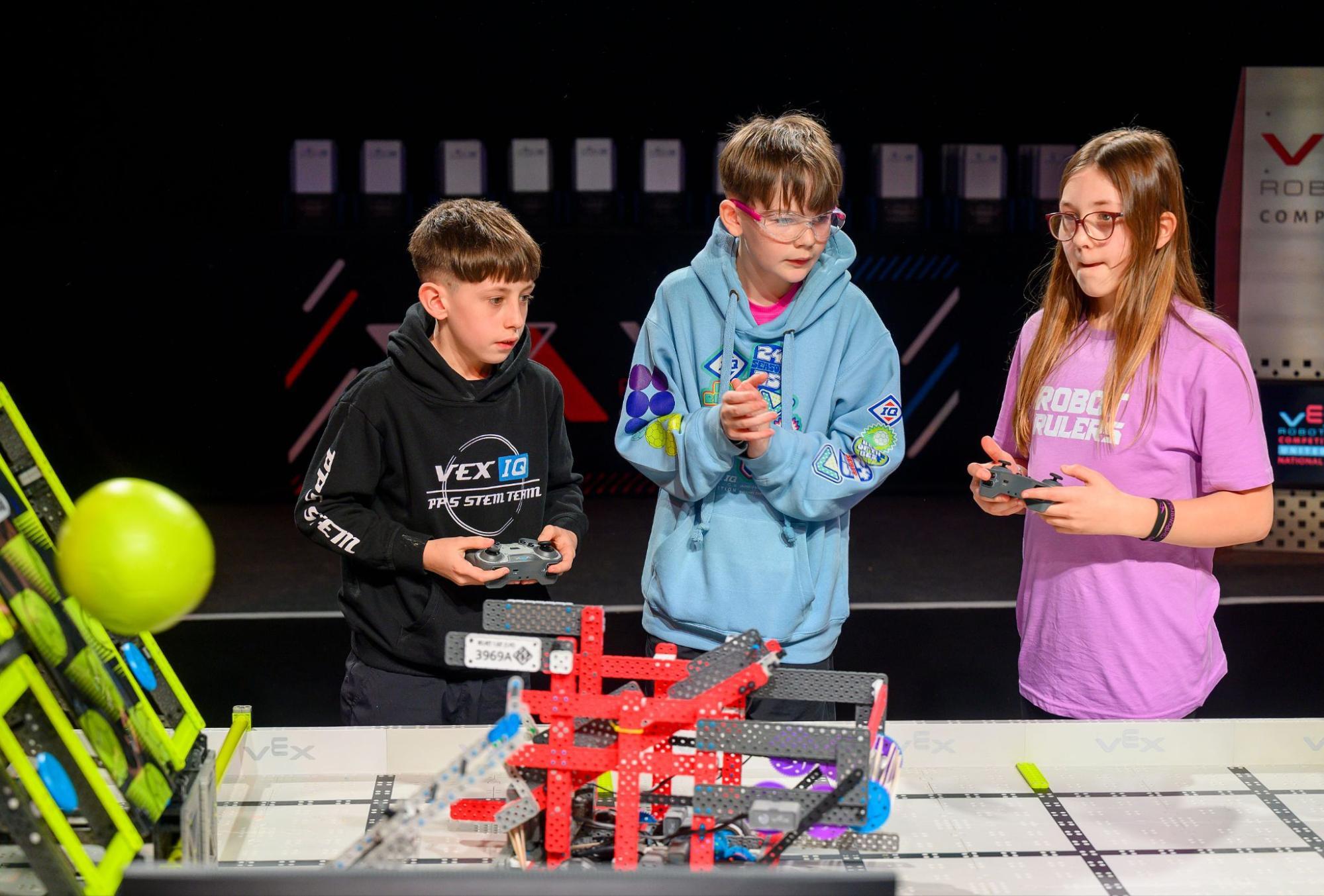 Two drivers, one boy and one girl, drive their robots on the Field during a match with a loader ready beside the field.