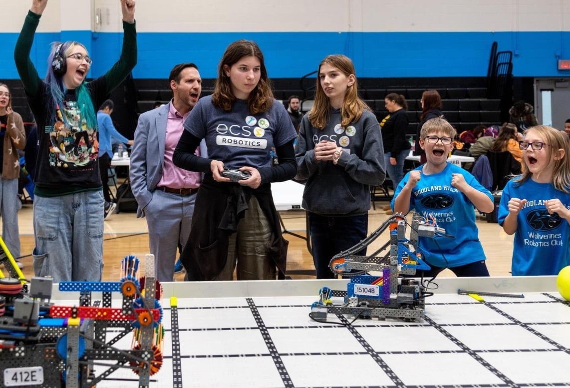 A student drives during a VEX IQ match while students from both teams and an adult cheer from beside the field.
