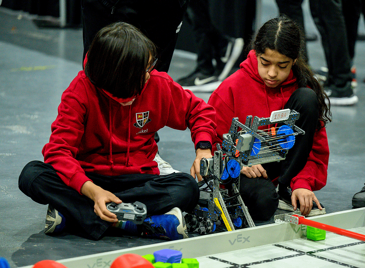 Two students on a team in matching shirts sit beside a game Field, with their robot between them as they work on it, with one holding a controller.