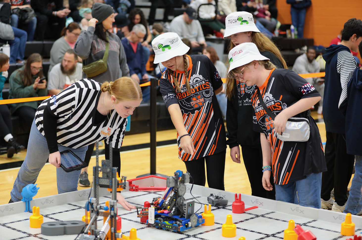 A referee holding a tablet leans over a VEX IQ field to score a match while three students point out where their robot finished.
