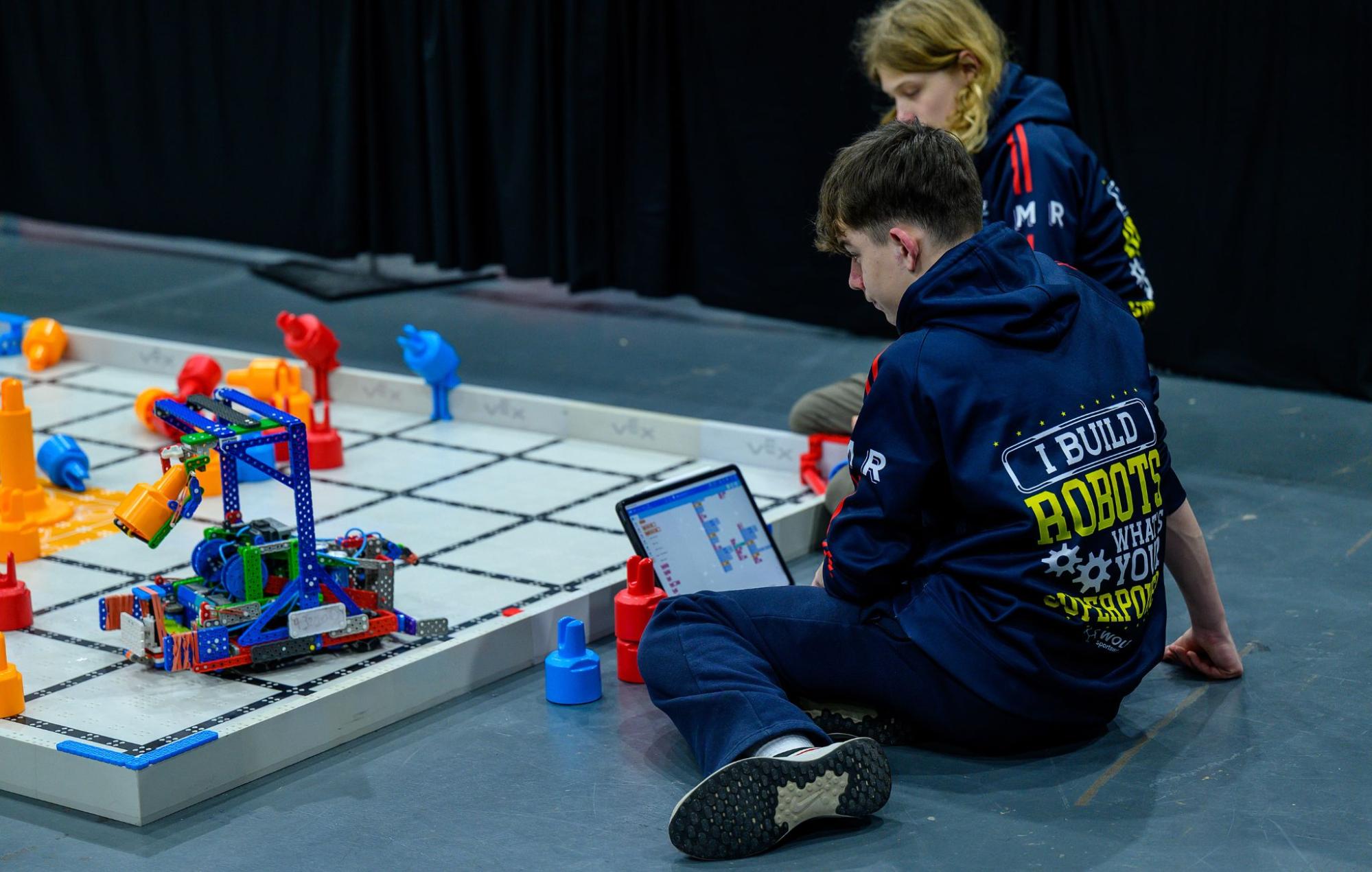 A student sits on the floor beside a VEX IQ field with a tablet, editing a block coding project, while a teammate works at the field behind them.