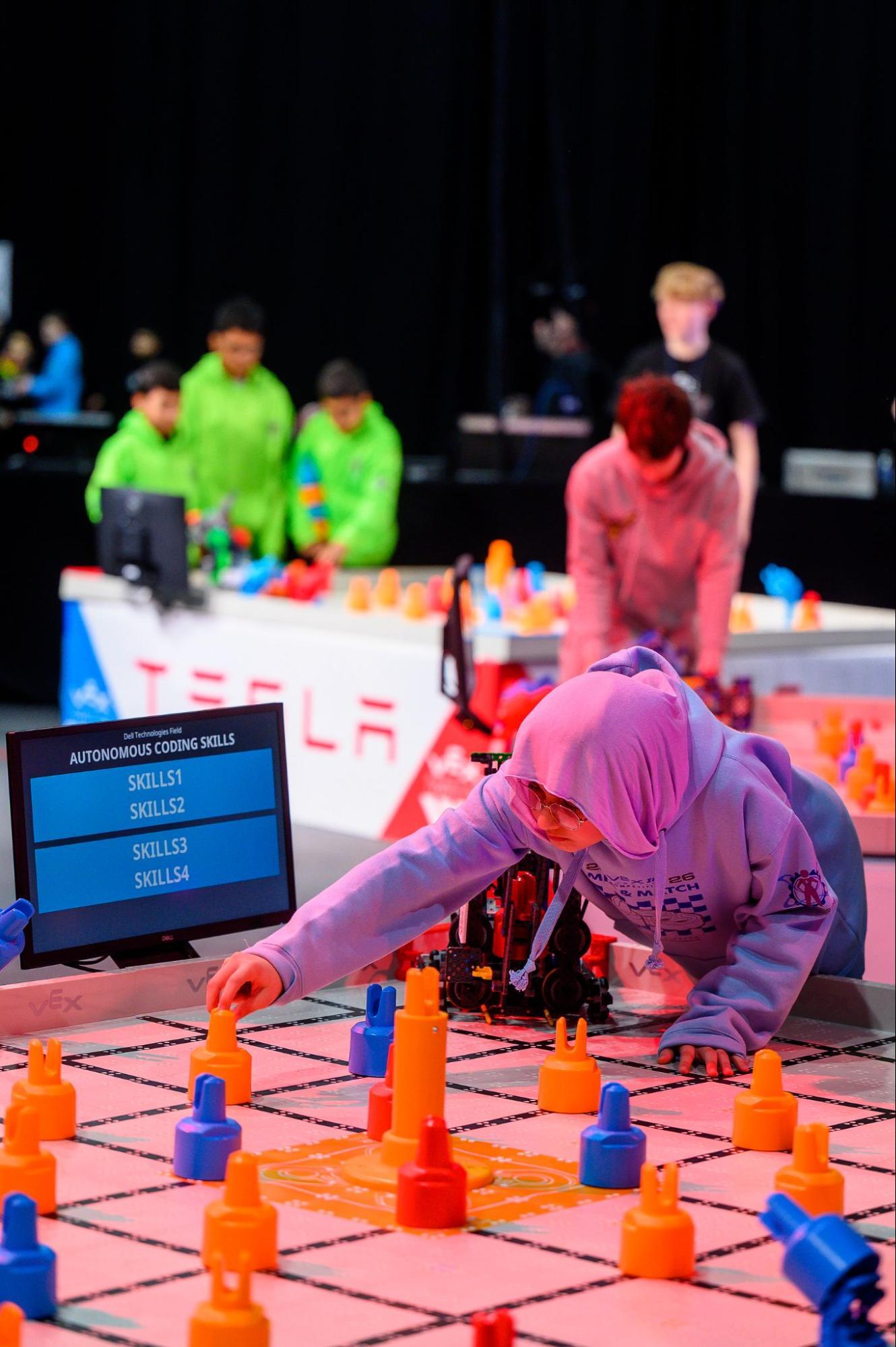A student reaches across a field to put a piece back in place. Her robot is behind her on the field and a screen can be seen to the side of the field reading Autonomous Coding Skills.