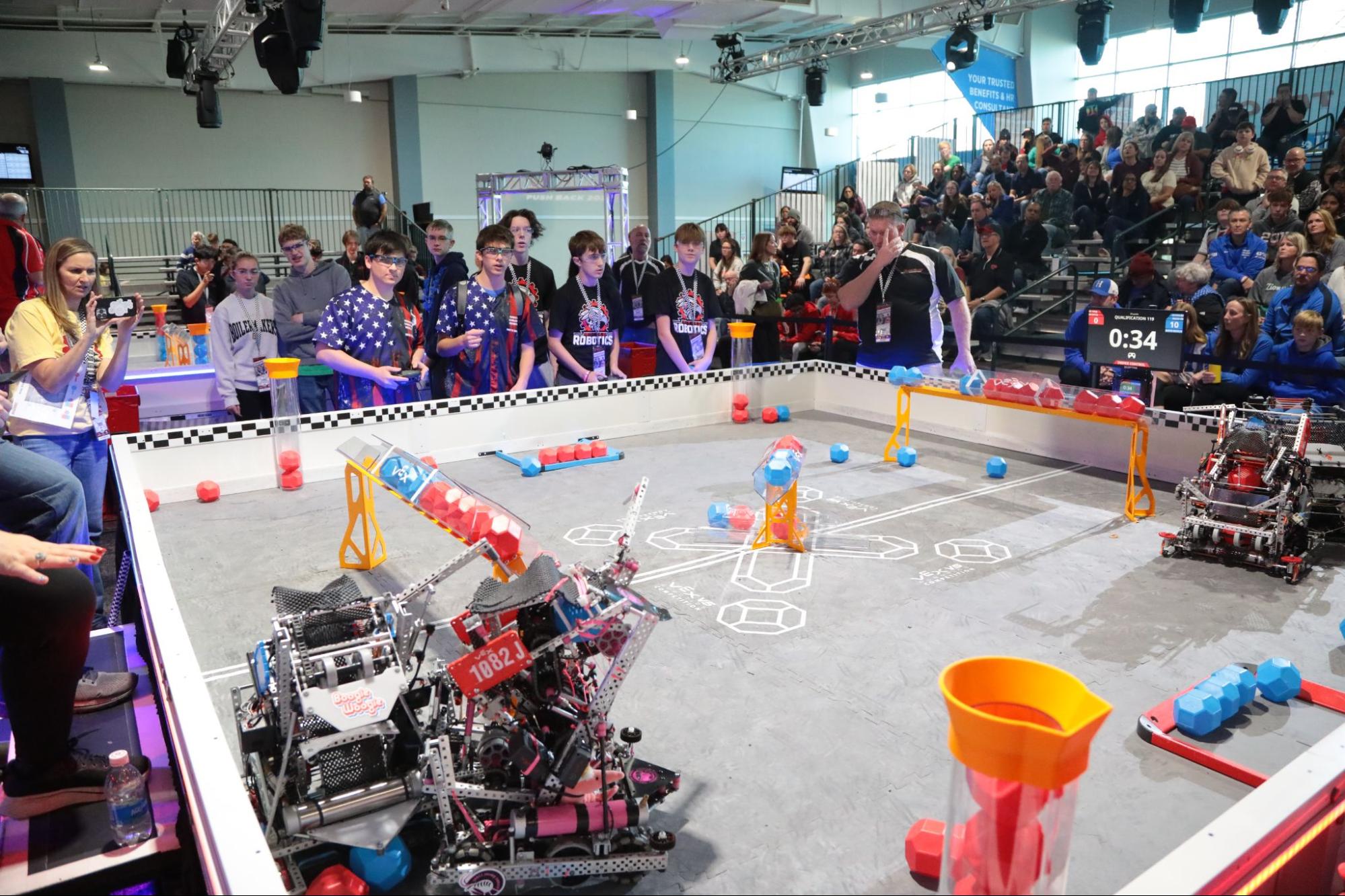 A VEX V5 match in progress with 34 seconds on the clock, two robots on the field, drive teams standing behind the field wall, and a full set of bleachers watching.