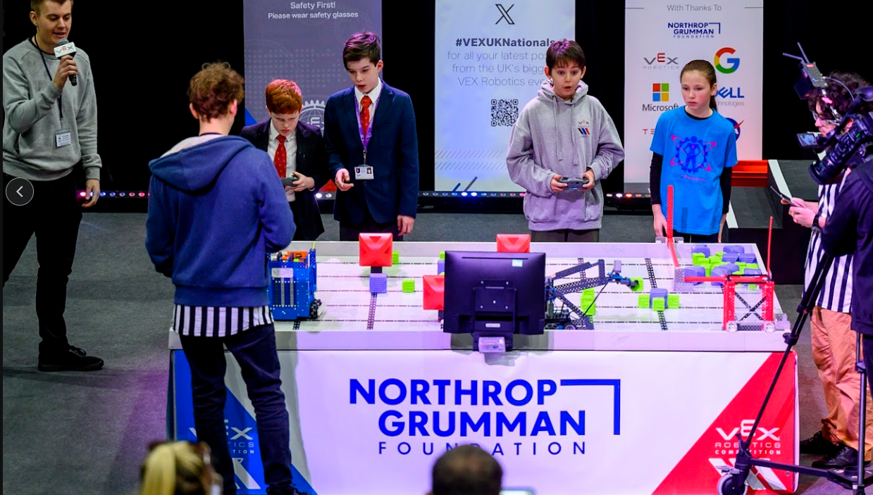 An emcee with a microphone speaks with a team beside a VEX IQ field while a camera operator films the match.
